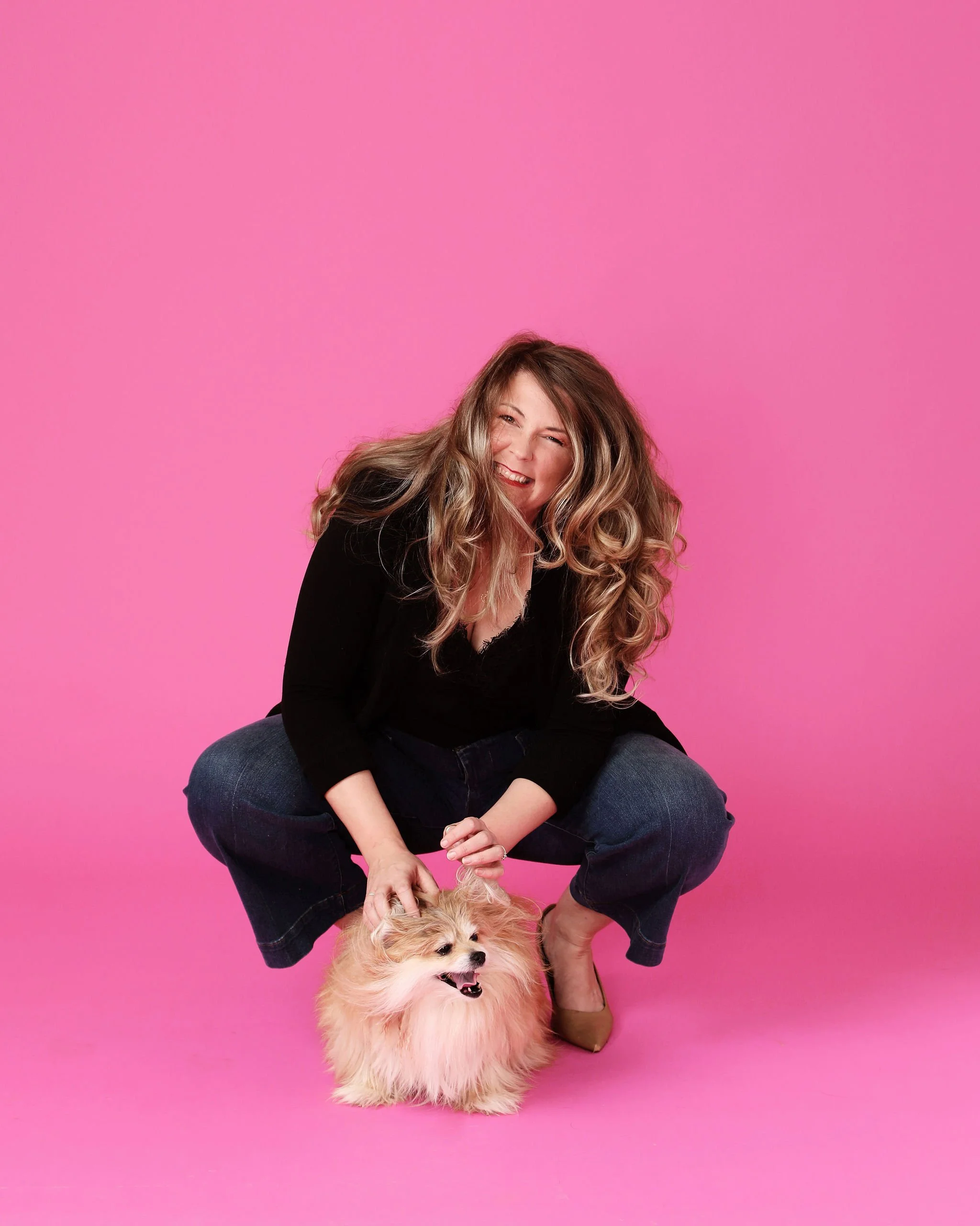 A woman with long wavy hair crouching down next to a fluffy small dog on a pink background.