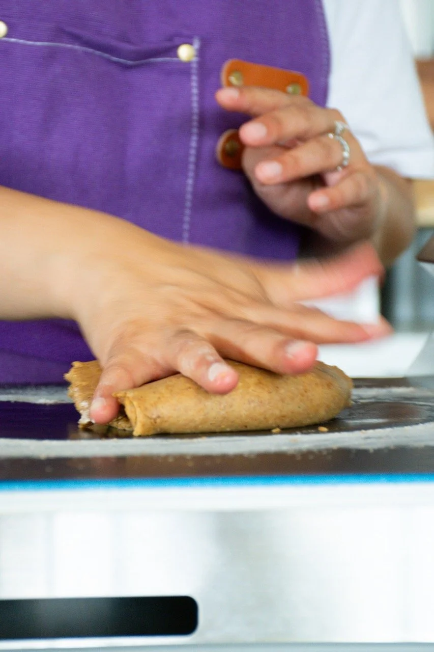 Person rolling a spring roll on a stovetop with a hand holding a phone nearby.