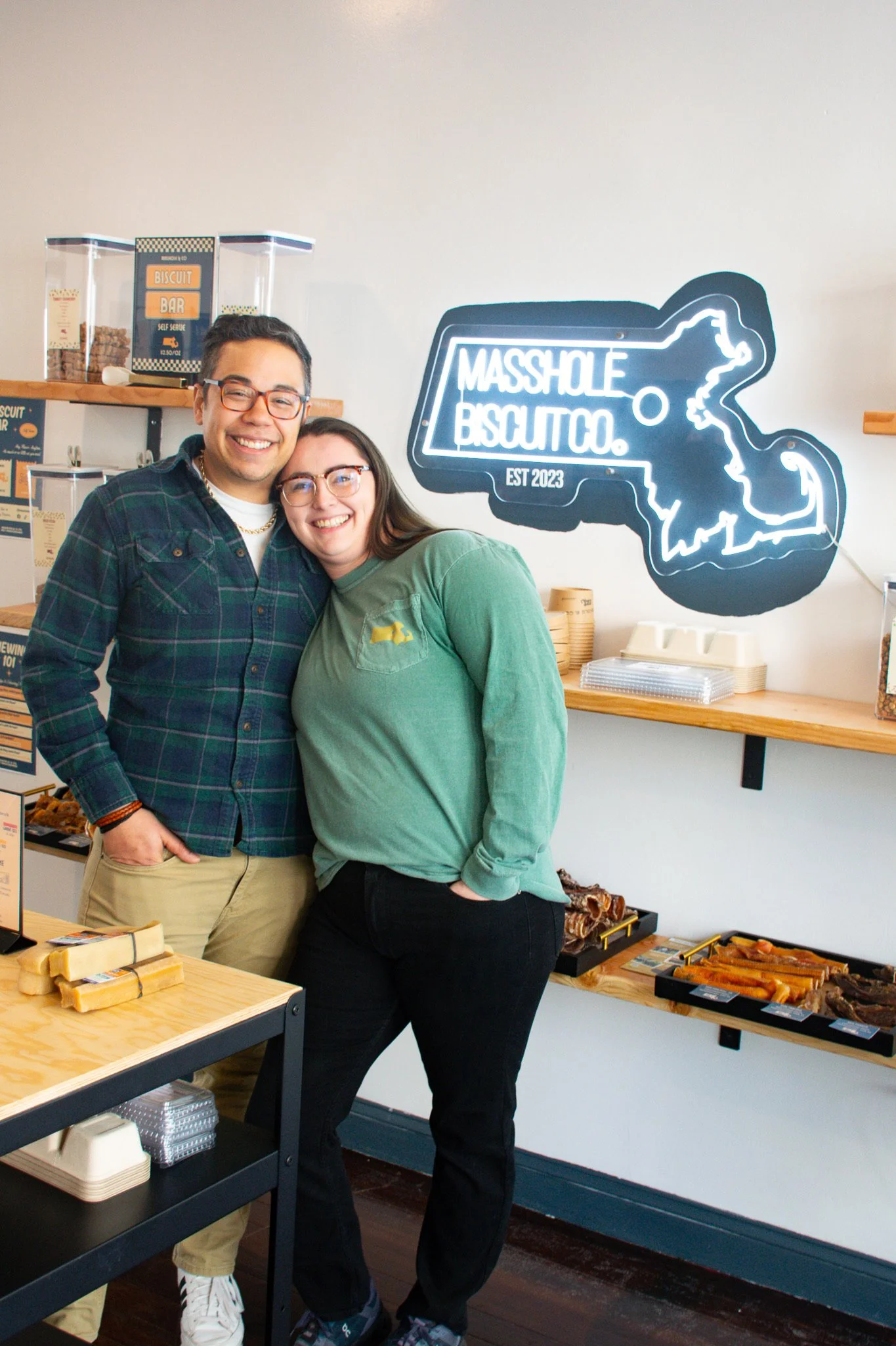 A smiling man and woman stand together inside a bakery named Mass Hole Biscuit Co., with a neon sign of the state of Massachusetts in the background. There are baked goods on the shelves and a wooden table with snacks in front of them.