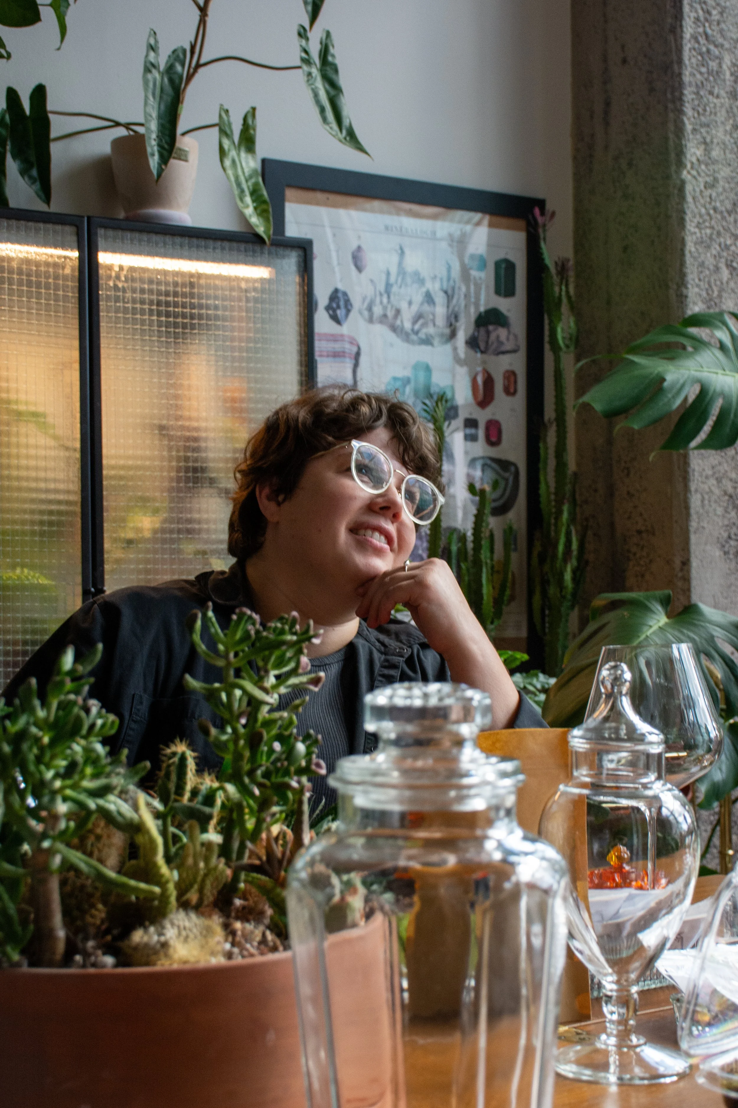 A person with short curly hair and glasses sitting at a table among potted plants and glass jars, smiling and looking off to the side.