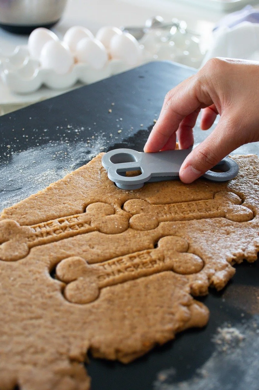 A hand using a bone-shaped cookie cutter to cut out cookies from rolled cookie dough on a black countertop, with a tray of eggs in the background.