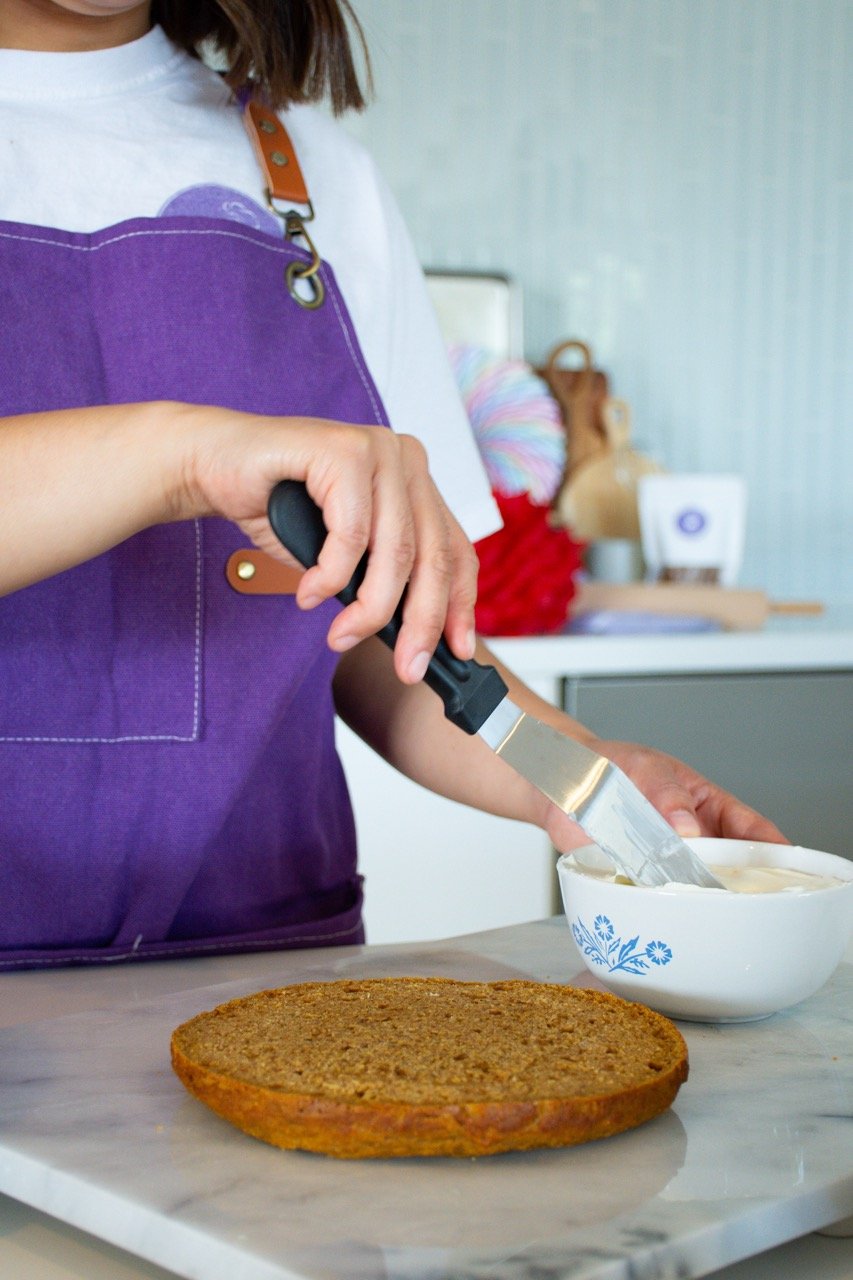 Person spreading cream cheese or butter on a graham cracker with a butter knife, wearing a purple apron in a kitchen.