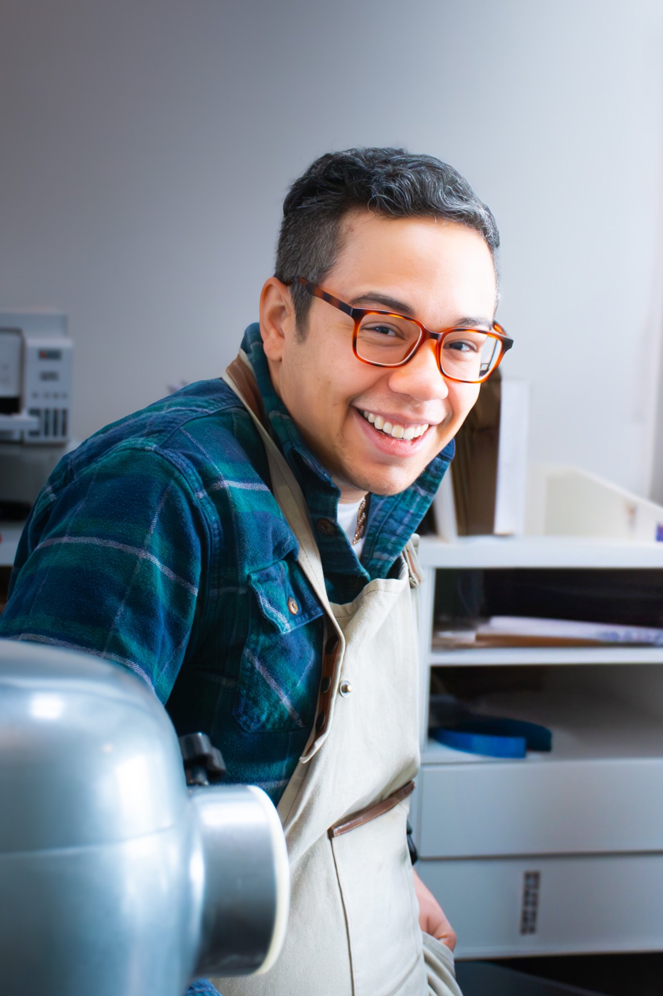 Noel is wearing a flanel shirt, with a brown apron on top. He smiles at the camera, standing behind a giant stand mixer.