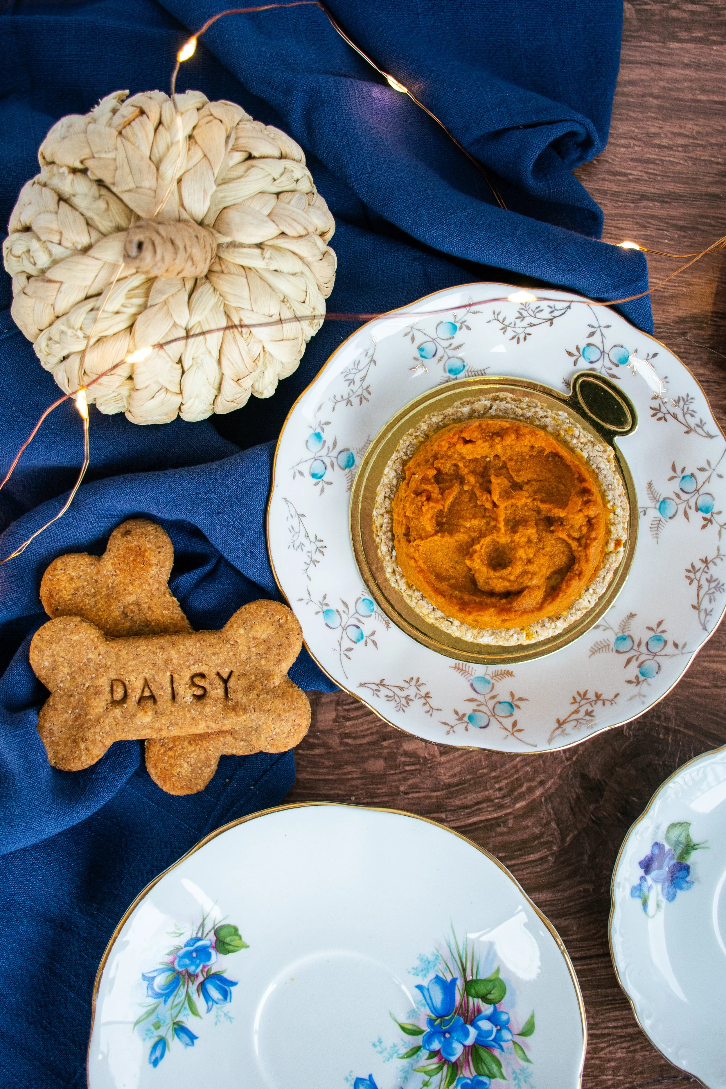 A pumpkin-shaped treat, two dog bone-shaped cookies with the name Daisy on one, a decorative pumpkin, and a pumpkin puree dish on a fancy floral plate, with a dark blue napkin and string lights on a wooden table.