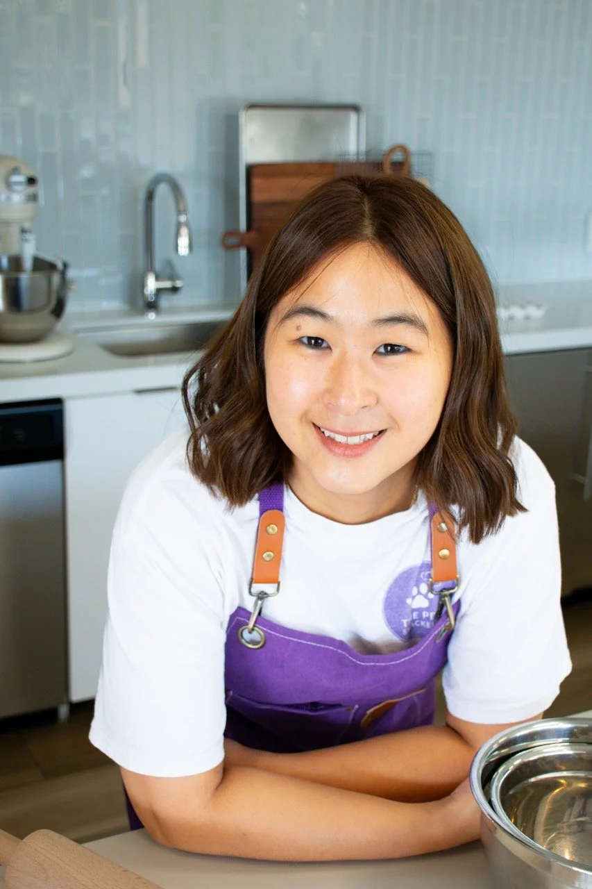 A woman with shoulder-length brown hair smiling in a kitchen, wearing a white shirt and a purple apron.