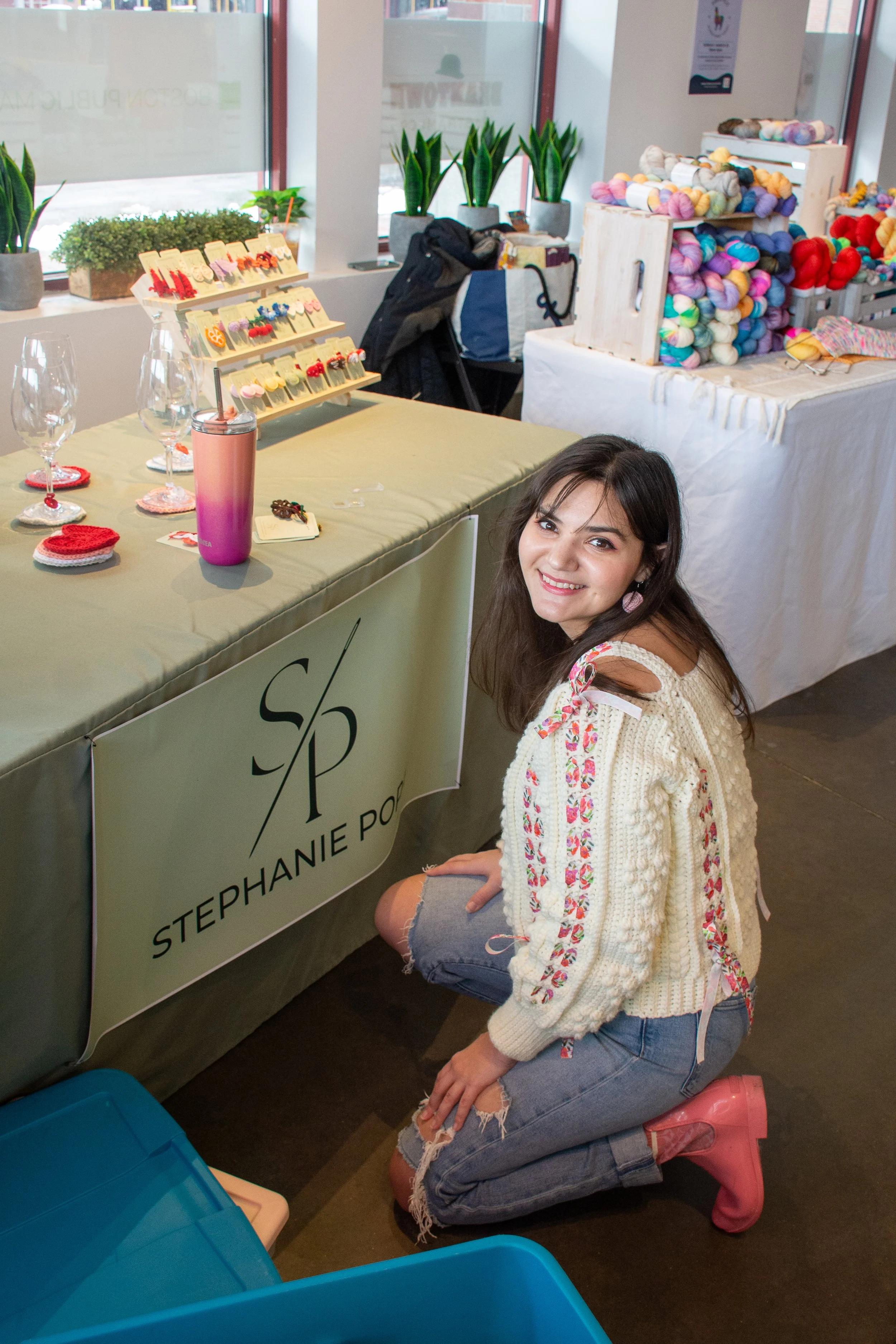 A woman kneeling beside a table with crochet crafts and jewelry, smiling at the camera, in front of a display of yarn at an indoor craft fair.