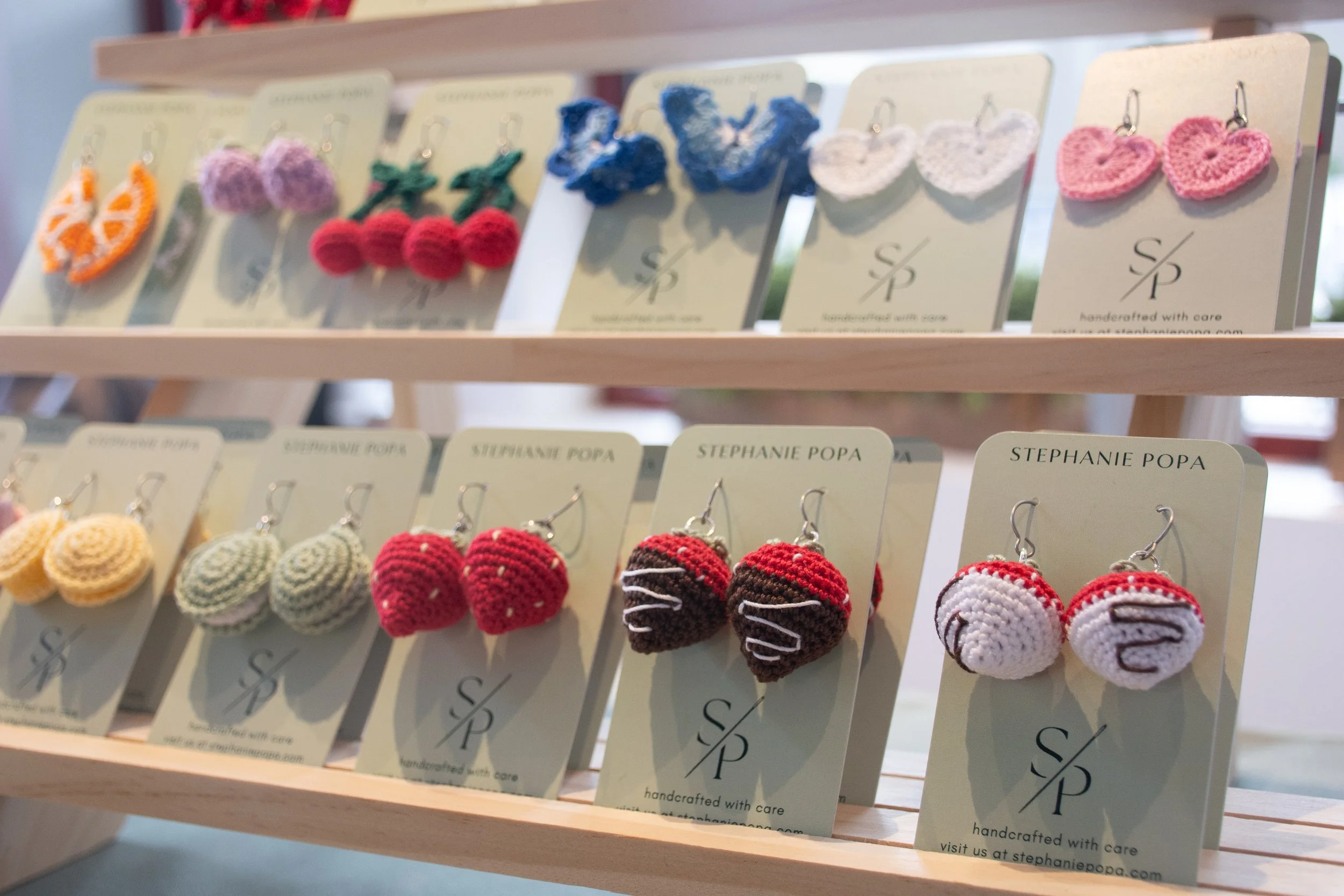 Display of handmade crocheted earrings on cards, arranged on wooden shelves at a craft stall.