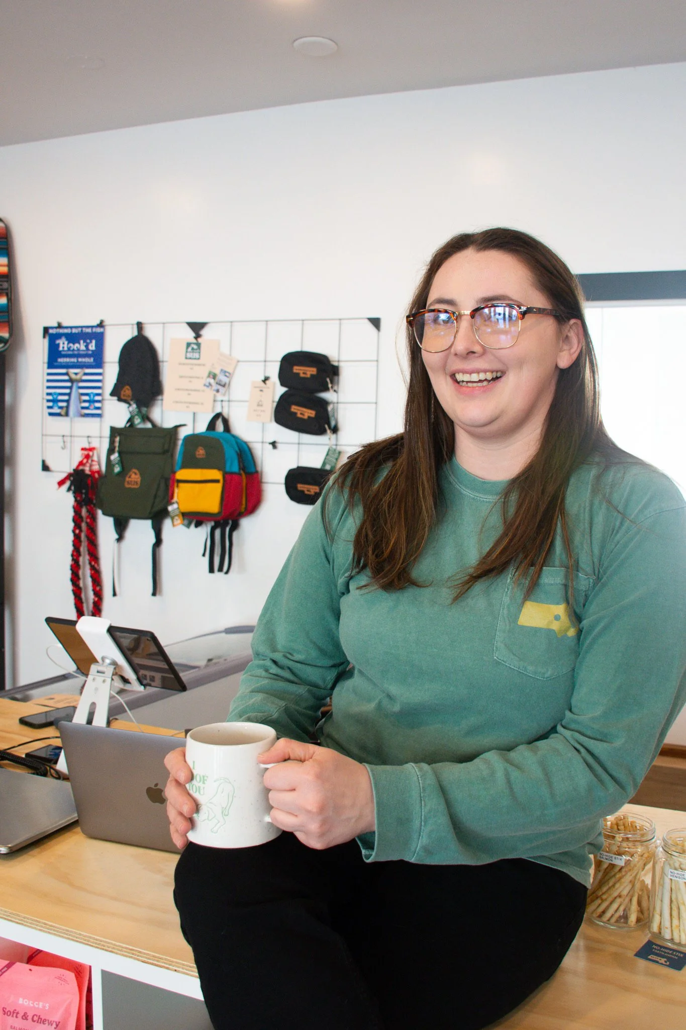 A woman with glasses holding a white coffee mug, sitting at a counter in a store or office. Behind her are backpacks and accessories on a wall display, with a laptop and electronic devices on the counter.