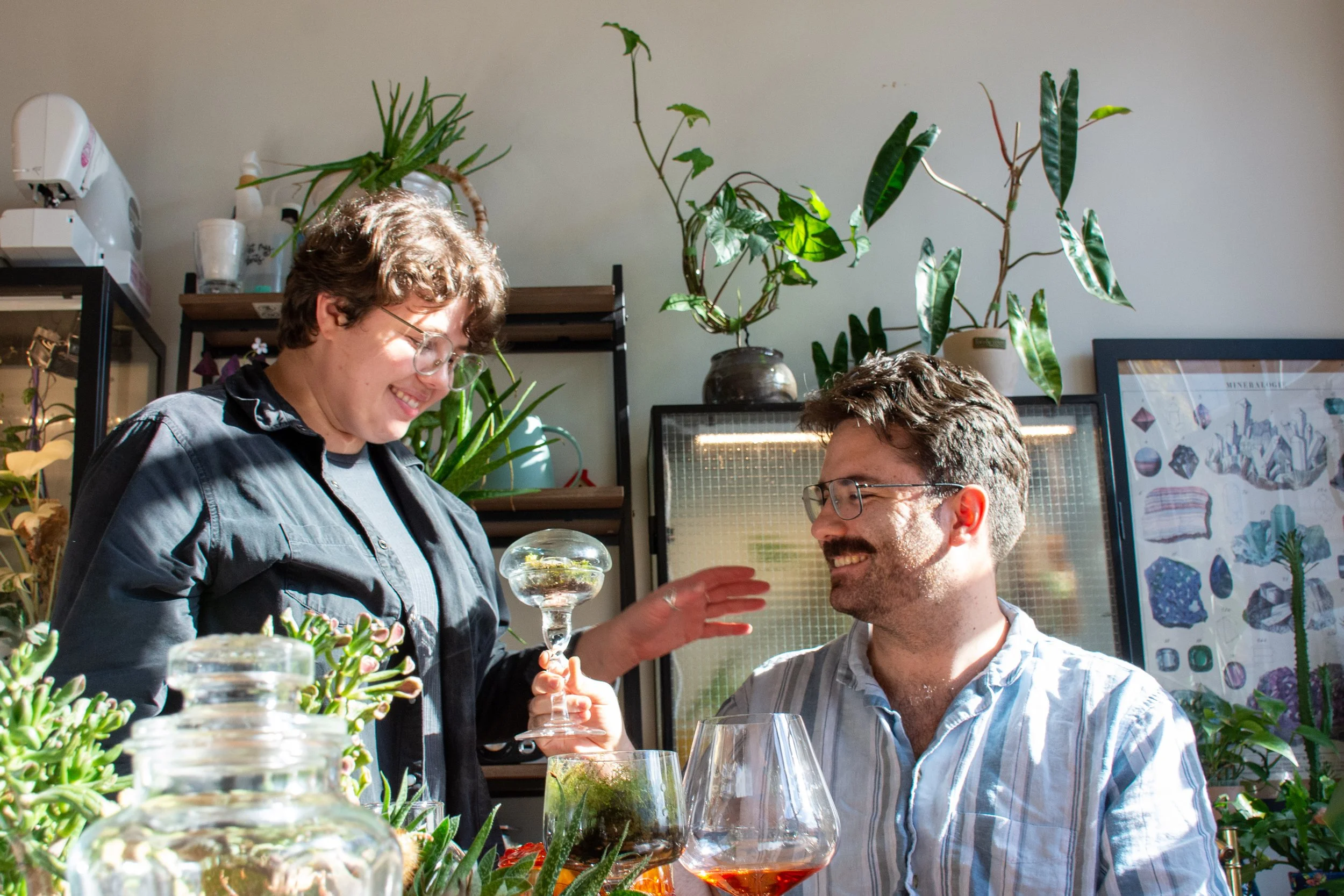 A woman and a man in a room filled with plants. The woman is holding a glass with a plant inside, smiling, and the man is sitting and looking at her, smiling. The room has sunlight and various decorative plants.