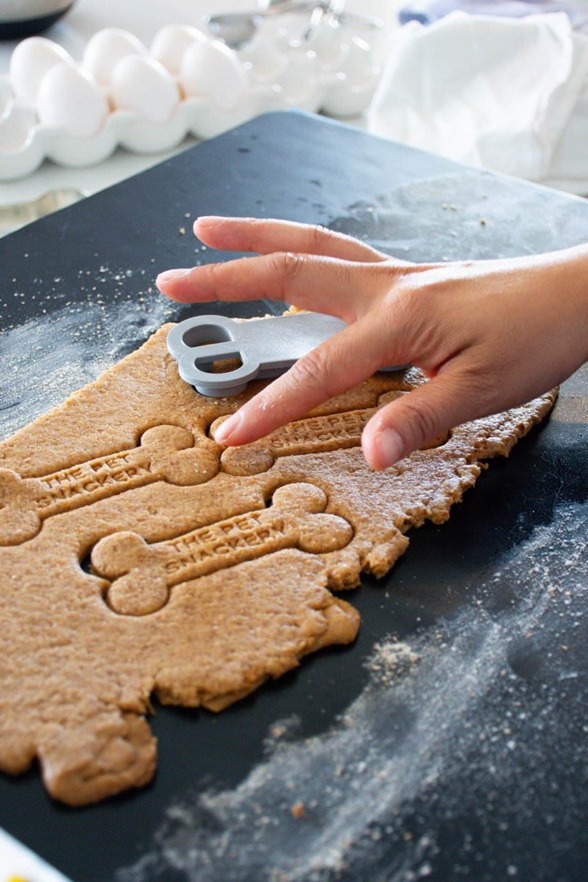 A person using a bone-shaped cookie cutter to cut out cookies from rolled dough on a black surface. Egg cartons and baking supplies are visible in the background.