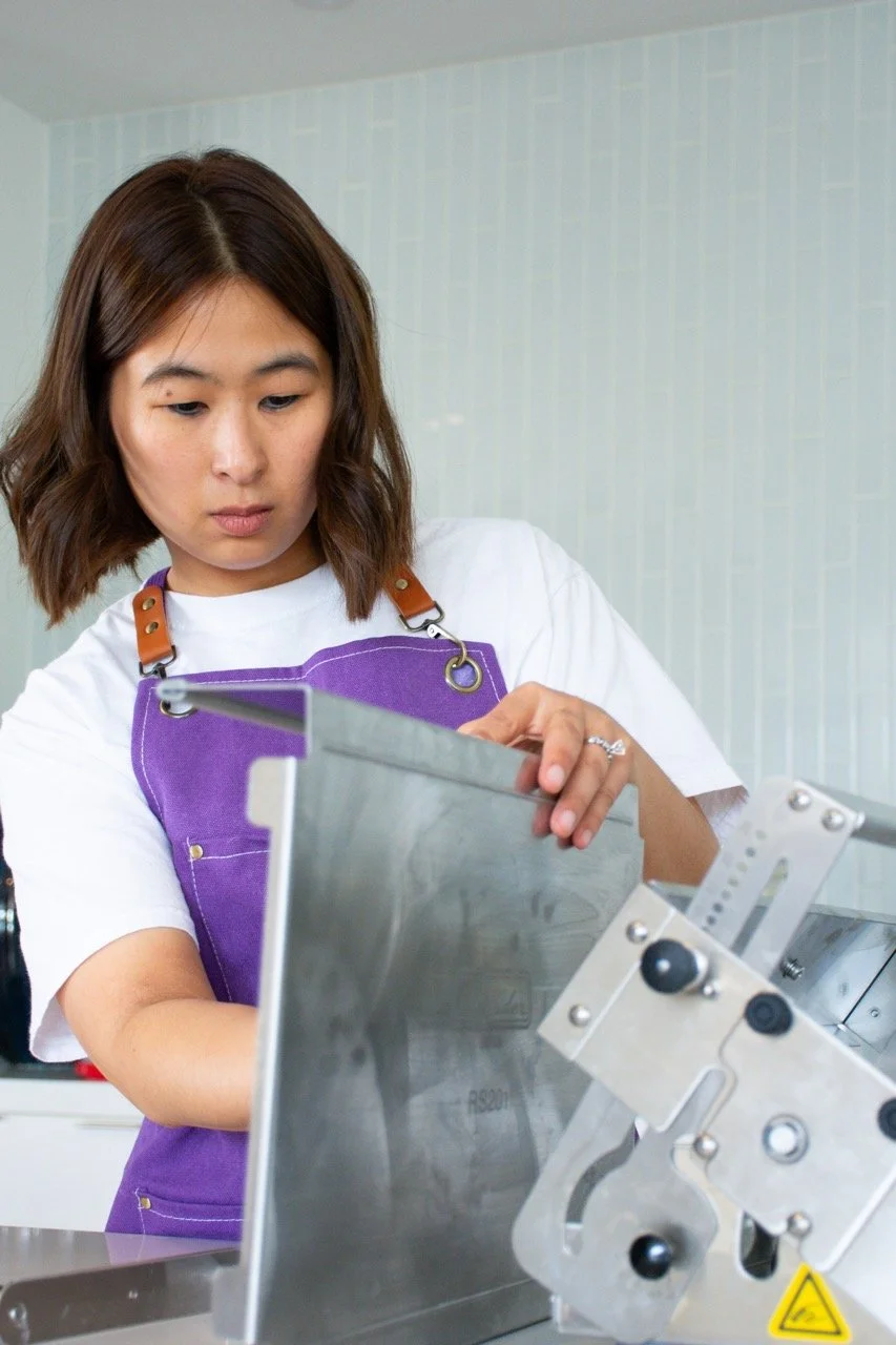 A young woman with shoulder-length brown hair wearing a white shirt and purple apron working with a metal machine in a clean workspace.