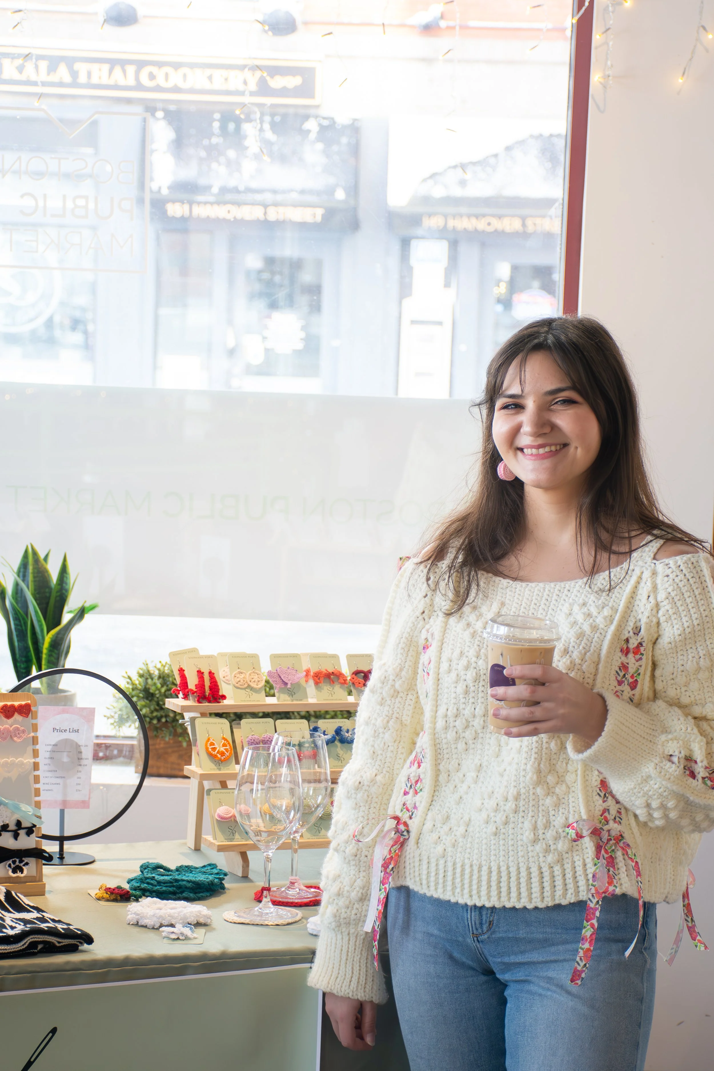 A woman smiling inside a store, holding a cup of coffee, with jewelry and accessories displayed on a table behind her.