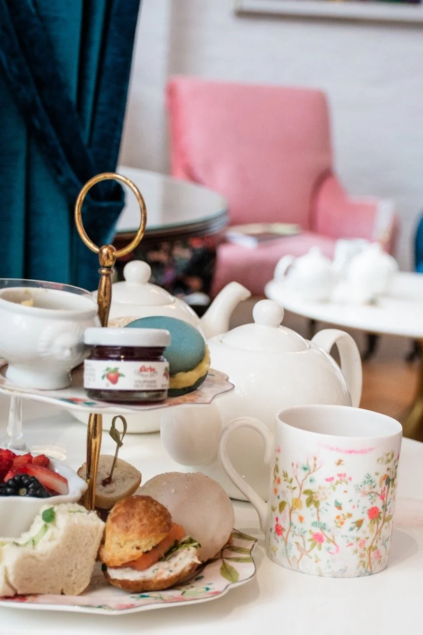 Tea set with teapot, teacup, and assorted pastries including scones, fruit tart, and macarons on a tiered serving tray, with a pink upholstered chair in the background.