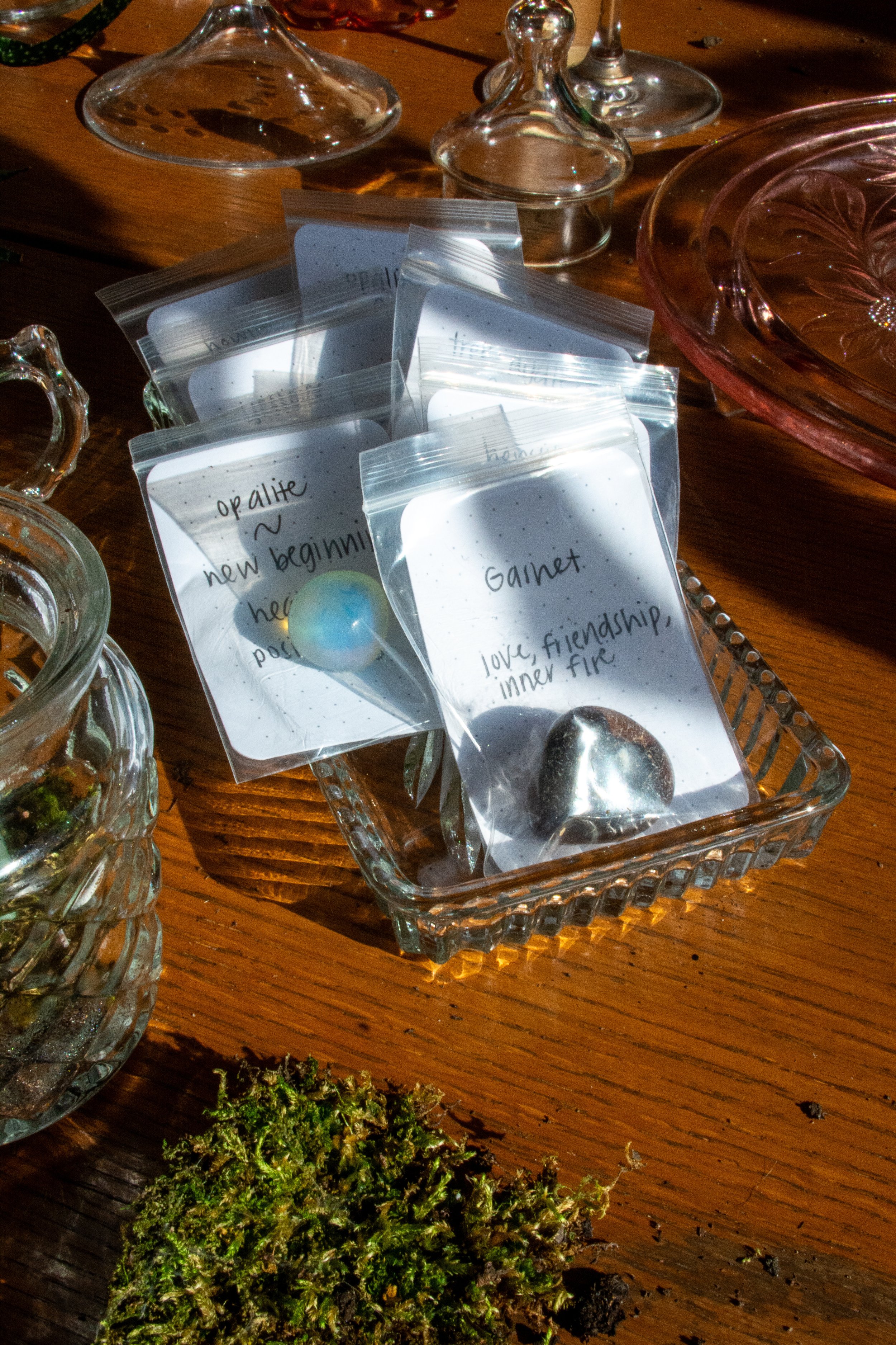 Small bags with handwritten notes and stones, surrounded by glass jars and a pink glass tray on a wooden table.