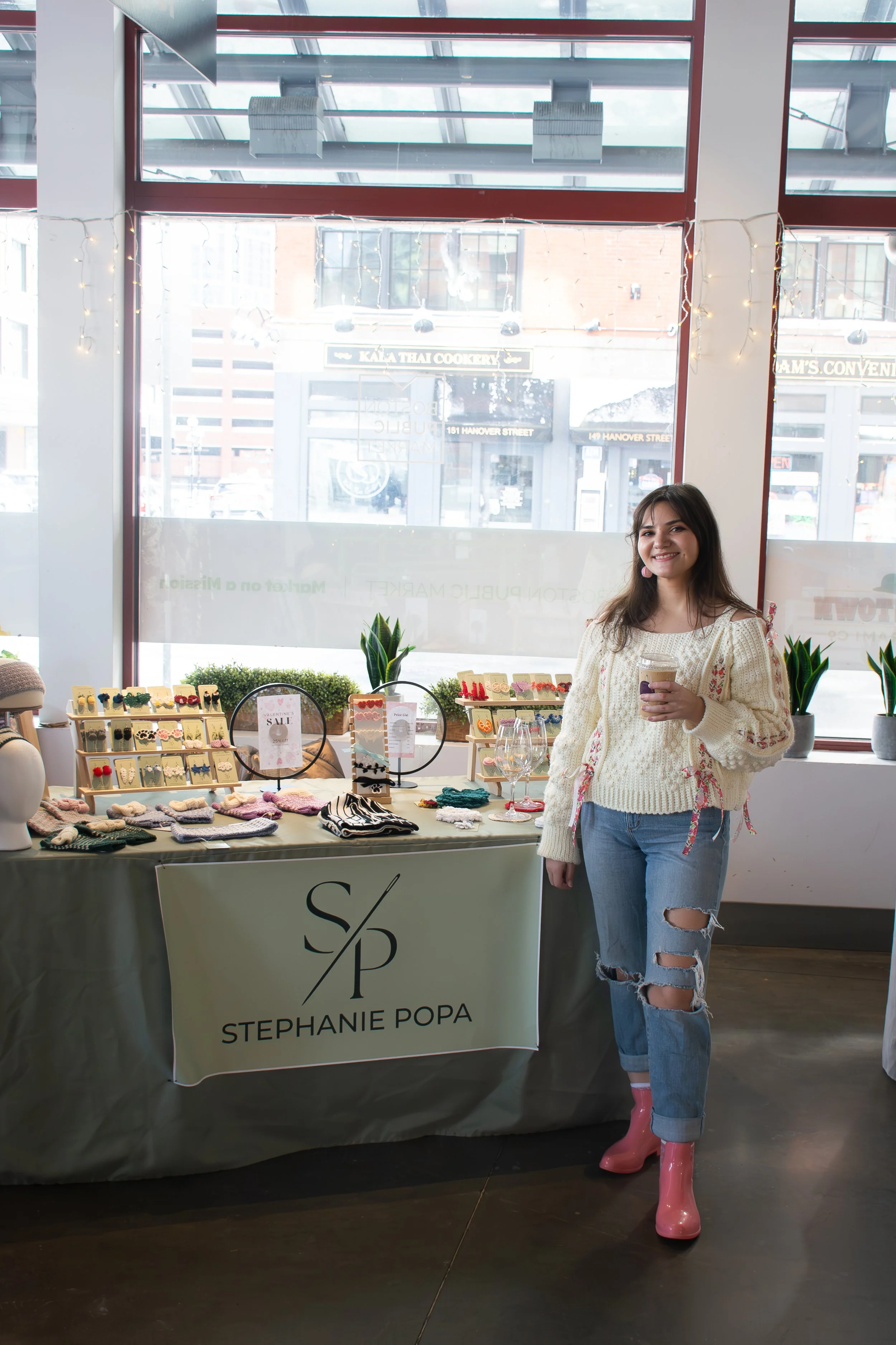 A woman standing next to a jewelry display table with a sign that reads 'Stephanie Popa'. She is smiling and holding a coffee drink, wearing a cream-colored sweater, ripped jeans, and pink rain boots inside a store with large window. Jewelry and acce