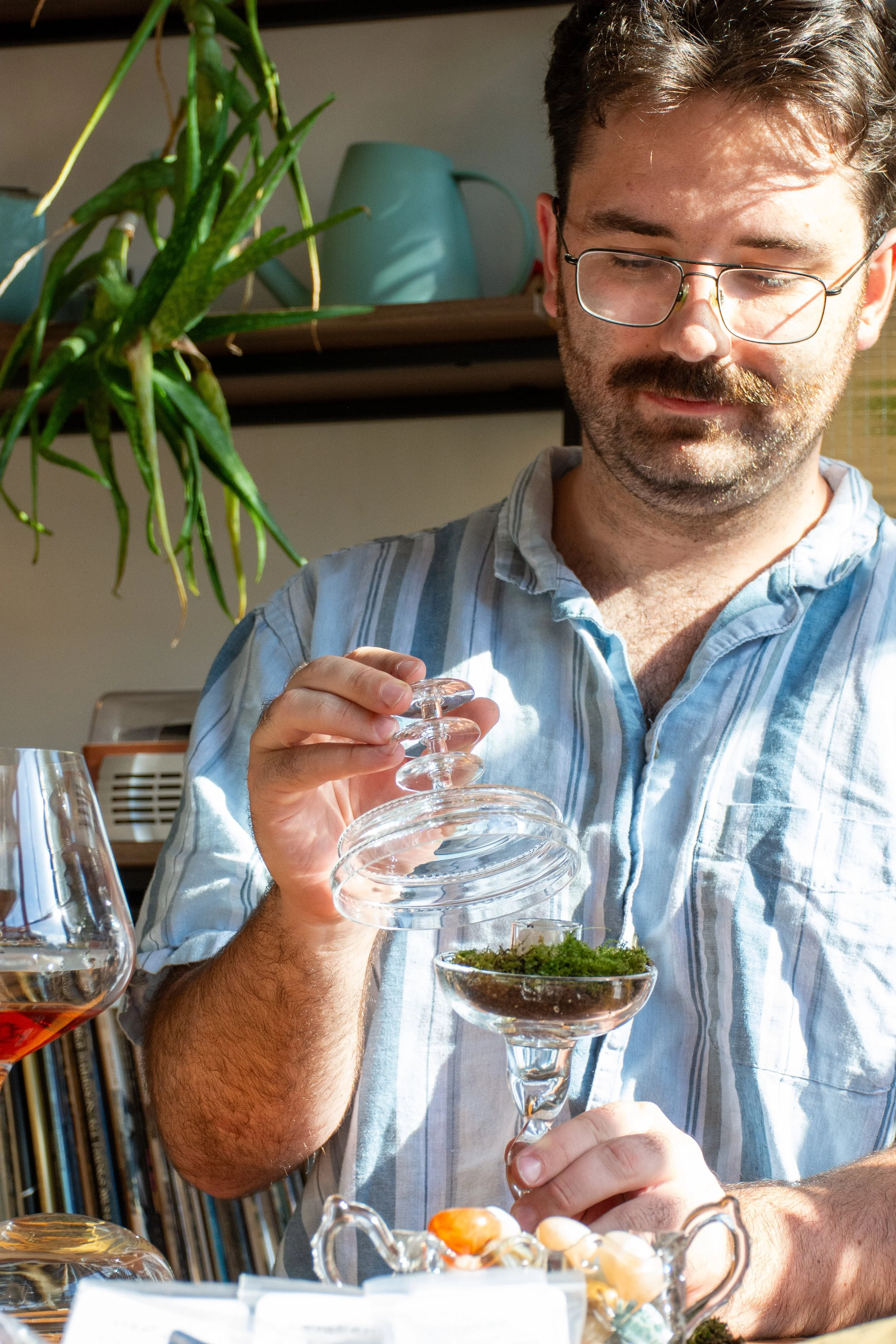 A man with glasses and a beard pours moss from a glass cloche into a martini glass.