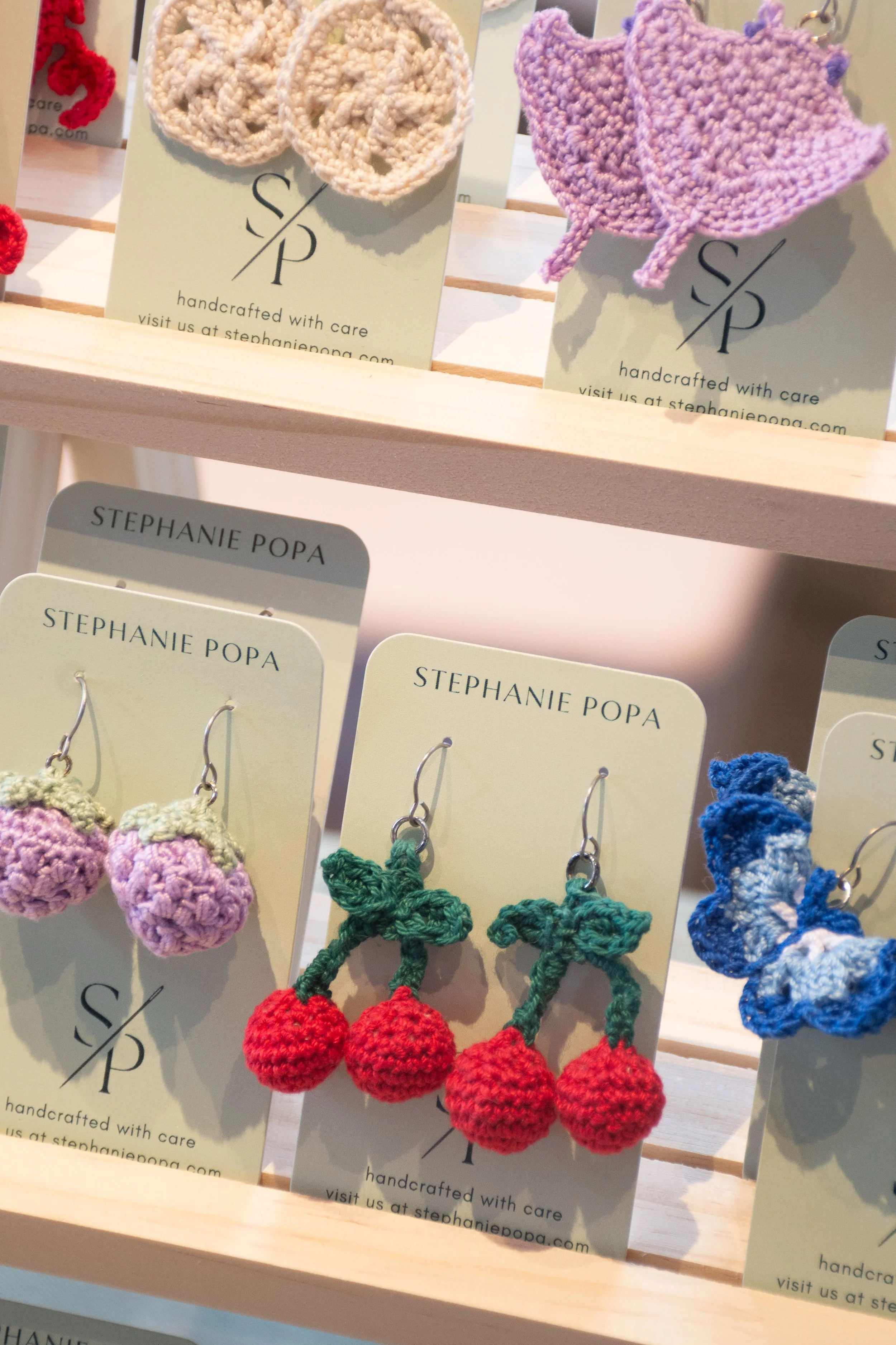 Assorted handcrafted crochet earrings shaped like strawberries, strawberries with green leaves, and light purple hats, displayed on a jewelry stand with tags from Stephanie Popa.