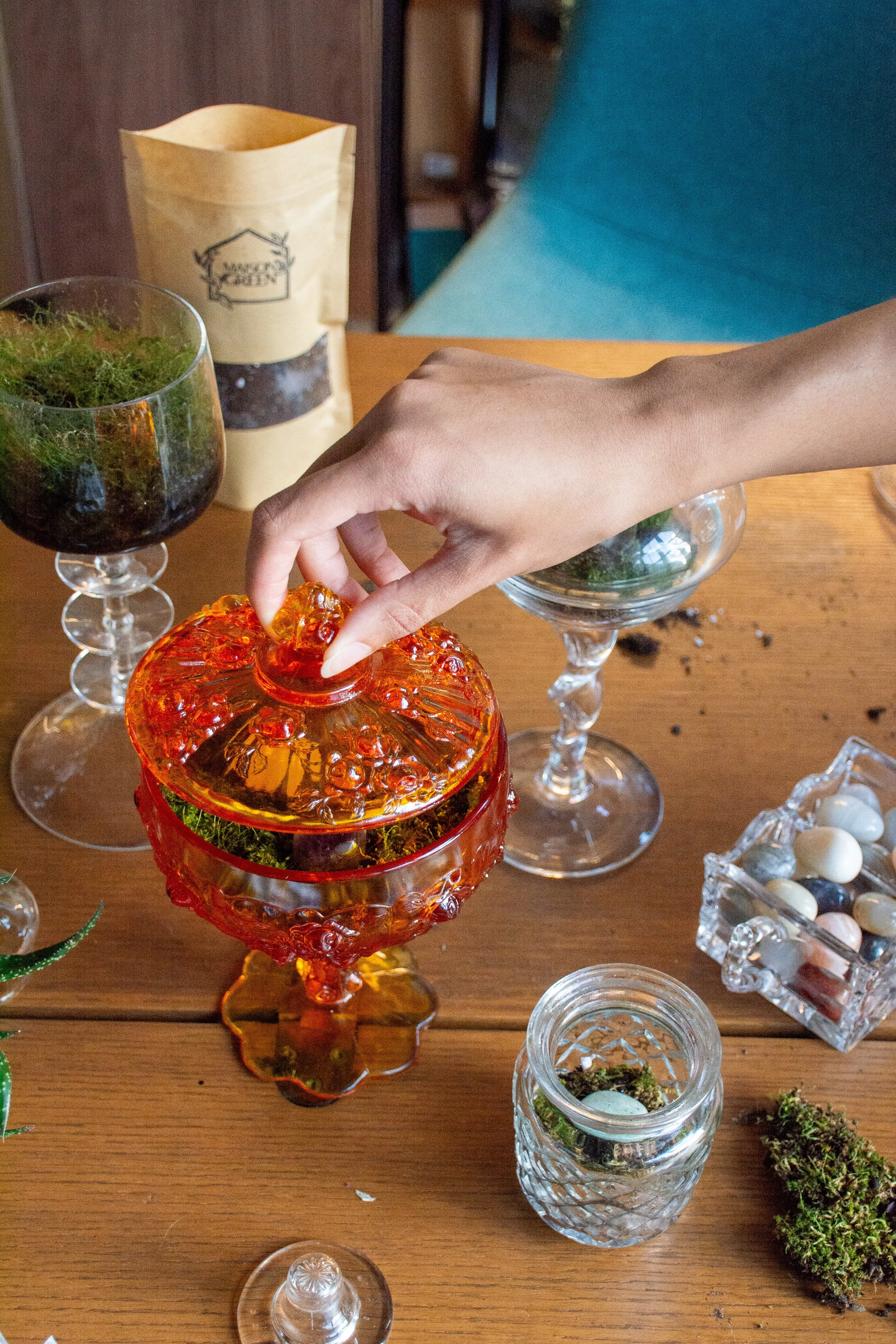A hand lifting the lid of an orange glass candy dish on a wooden table, surrounded by various glassware, a jar of dried herbs, and a small tray of marbles.