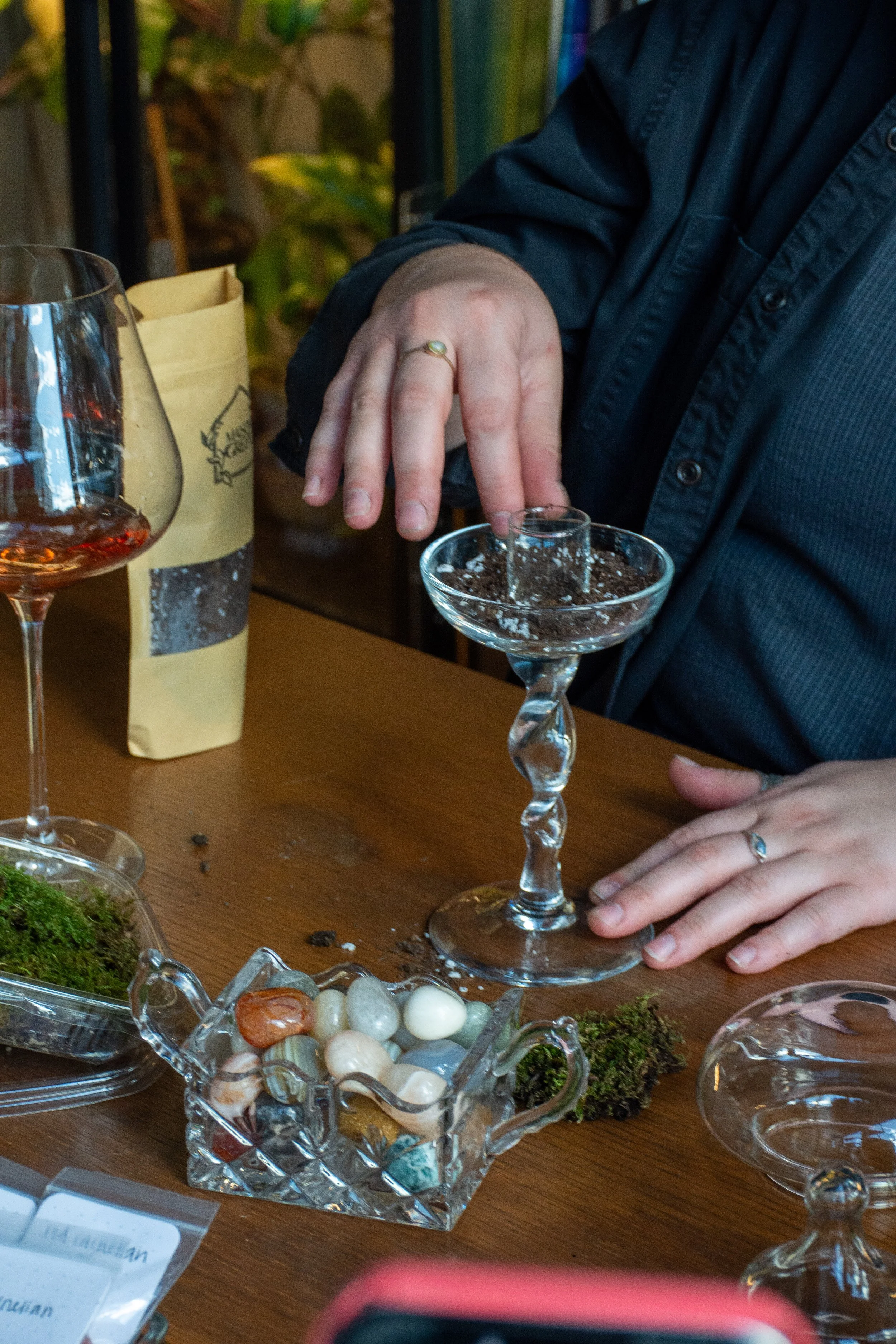 Person placing a small glass into a wider, twisted glass stemmed container on a wooden table with colorful stones, moss and wine glasses.