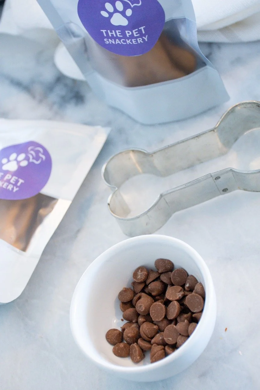 Chocolate pet treats in a white bowl, cookie cutter, and pet treat packaging on a marble surface.