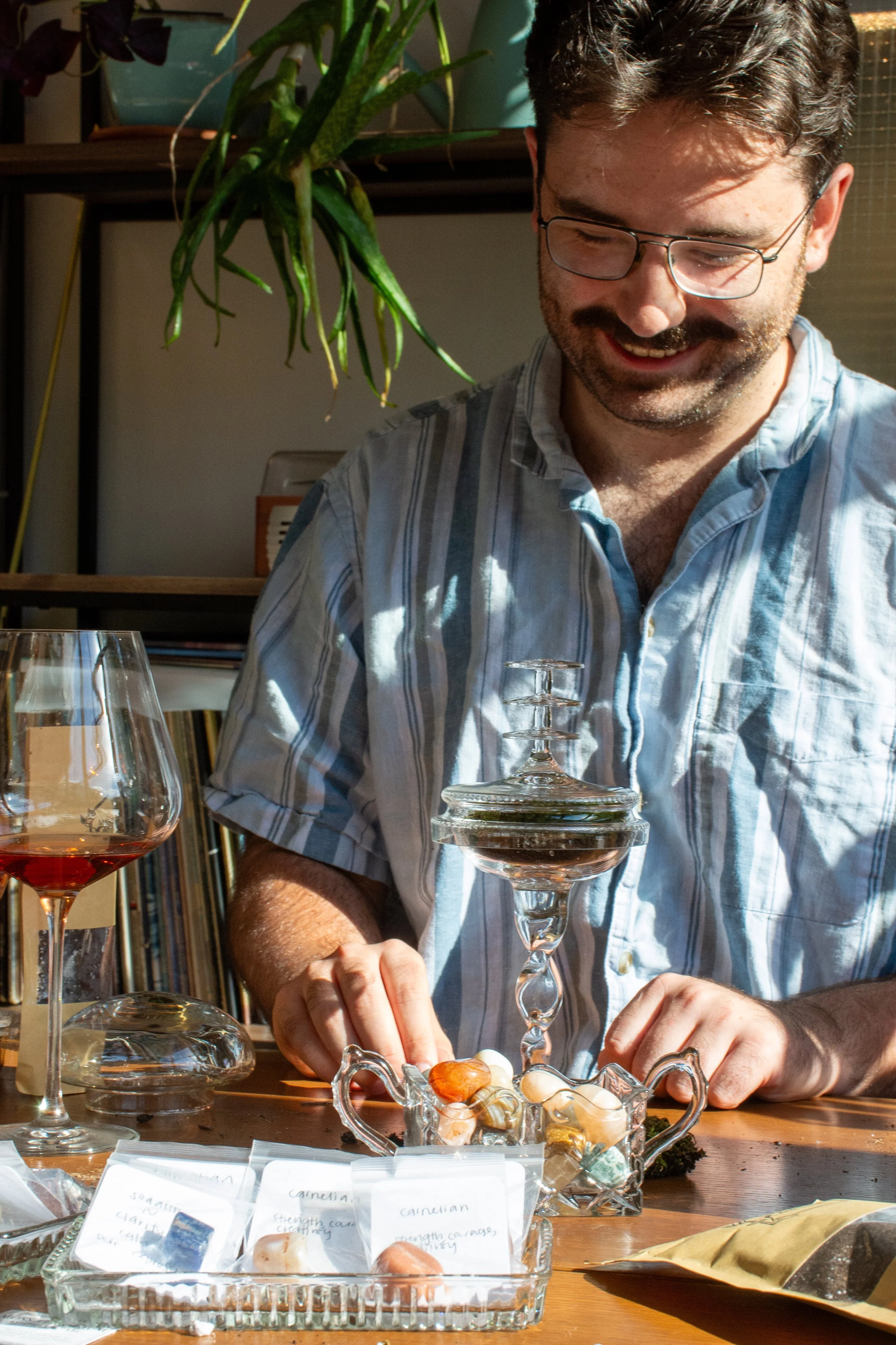 A man with dark hair, glasses, and a beard, smiling and looking down, sitting at a table with a glass of rosé wine, and what appears to be mineral or gemstone samples in a decorative glass container, surrounded by labels and jewelry, in a well-lit ro