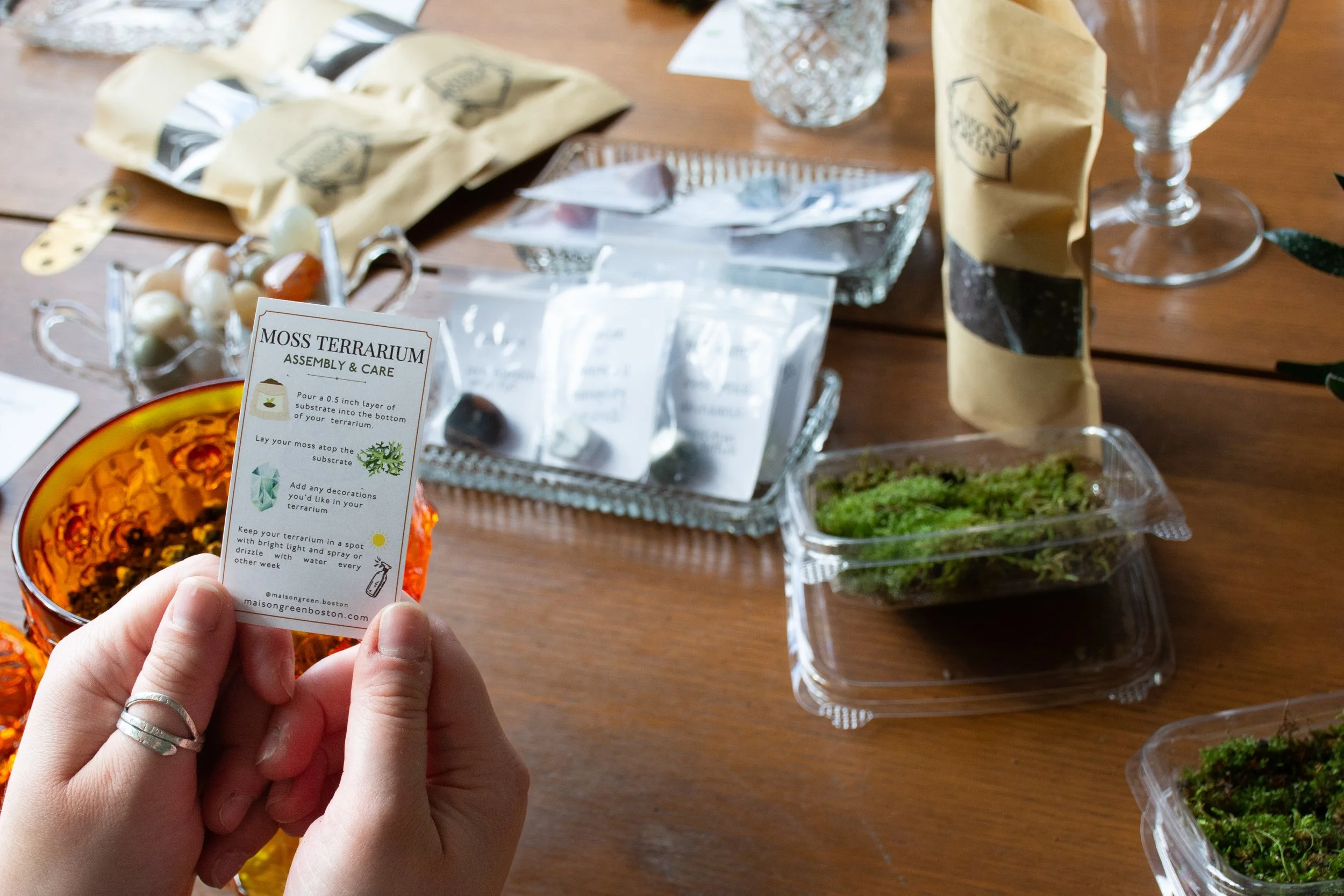 Person holding a moss terrarium assembly instruction card in front of a table with moss, decorative stones, and packaging materials for creating a moss terrarium.