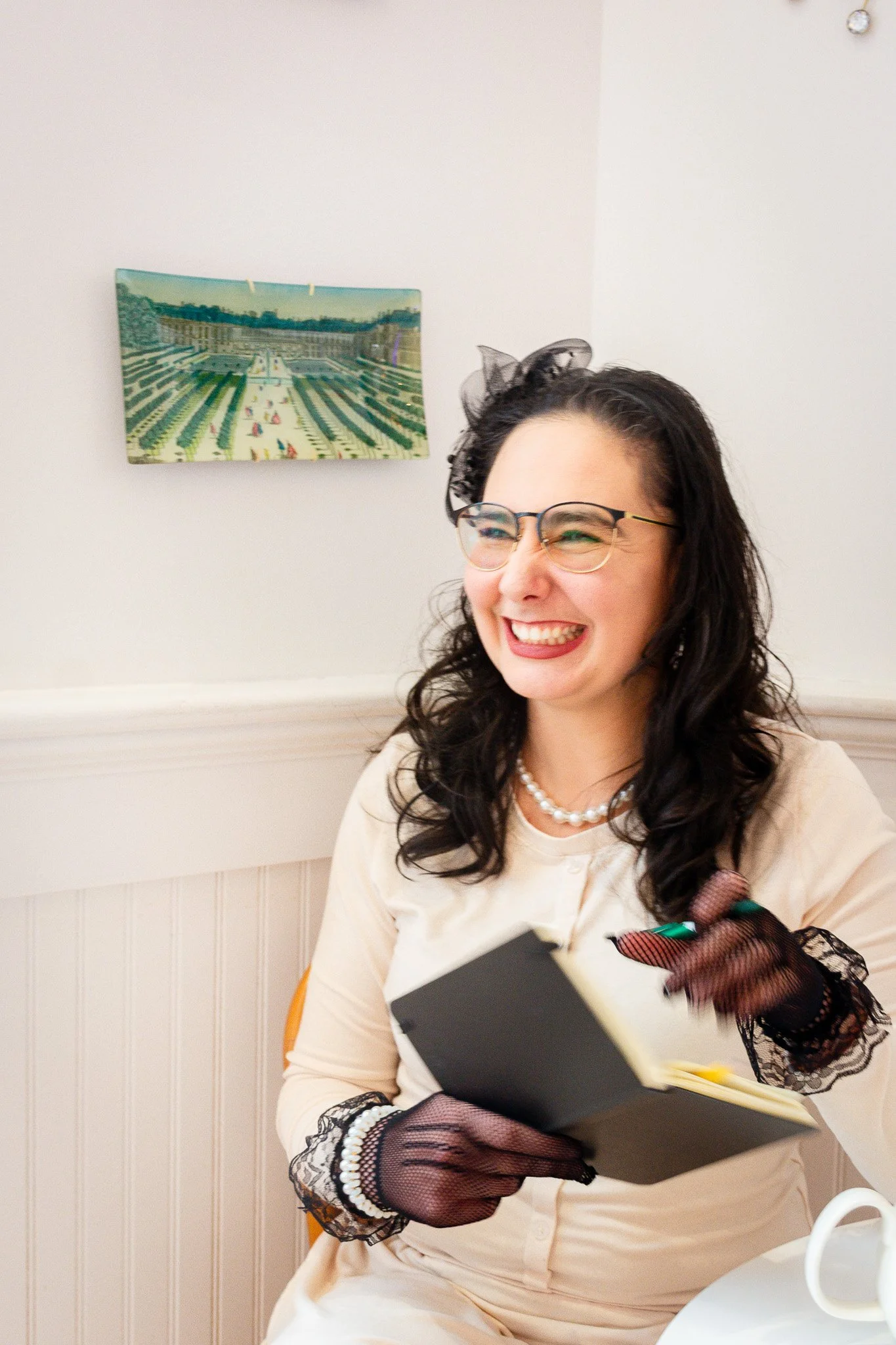 Amanda marie wears a cream colored sweater and glasses with a feather fascinator in her hair. She is holding a notebook and pen, looking off in the distance and smiling.