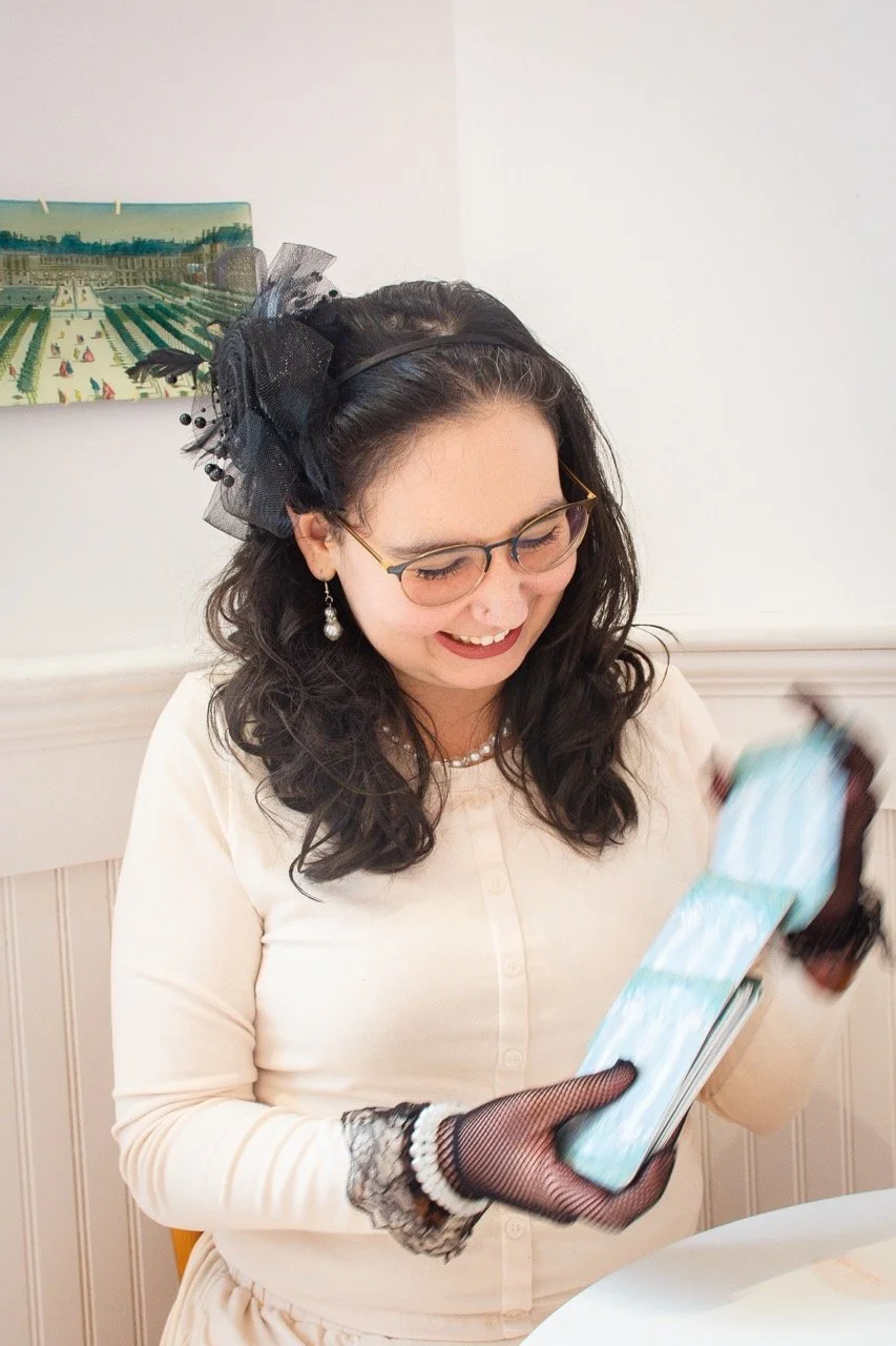 A woman with dark curly hair, glasses, and pearl earrings smiling while looking at her phone. She is dressed in a cream-colored sweater, wearing fingerless lace gloves, and has a black headband with a large black tulle bow. She is sitting at a table 