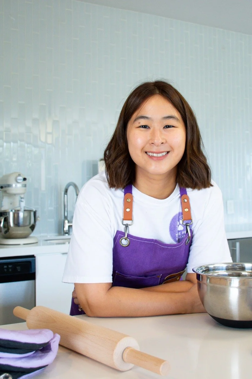 A young woman with shoulder-length brown hair smiling in a kitchen. She is wearing a white t-shirt and a purple apron, with a rolling pin on the counter in front of her and a mixing bowl to her right.