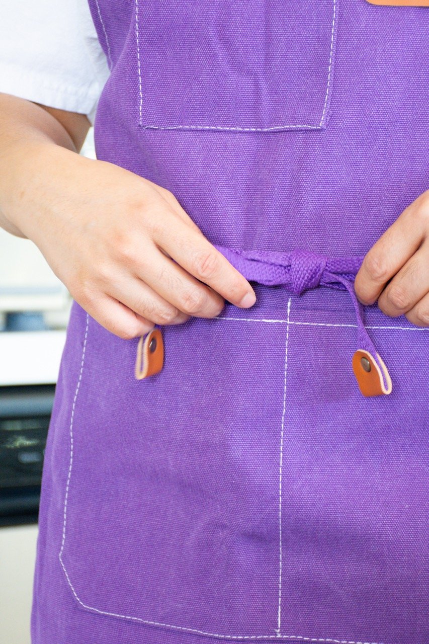 Person adjusting a purple apron with a front pocket and leather accents.