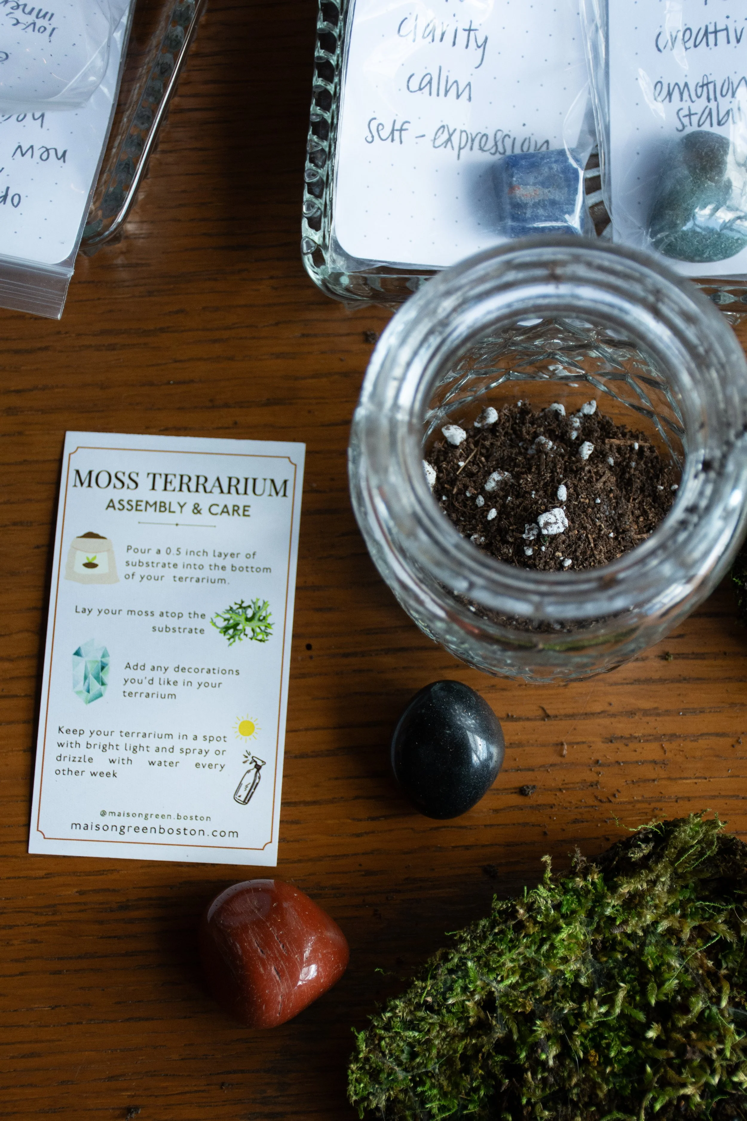 Moss terrarium making supplies including soil, decorative stones, moss, and instructions on how to assemble a terrarium.
