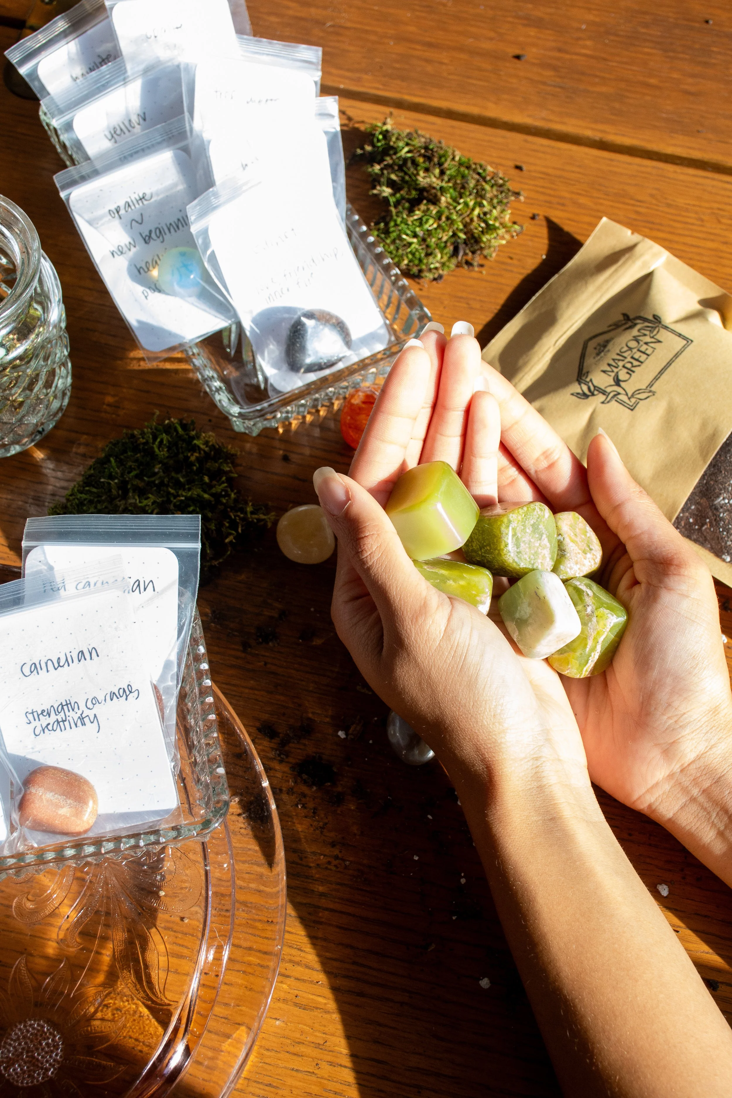 Person holding green and white gemstones, surrounded by various labeled stones and crystals on a wooden table.