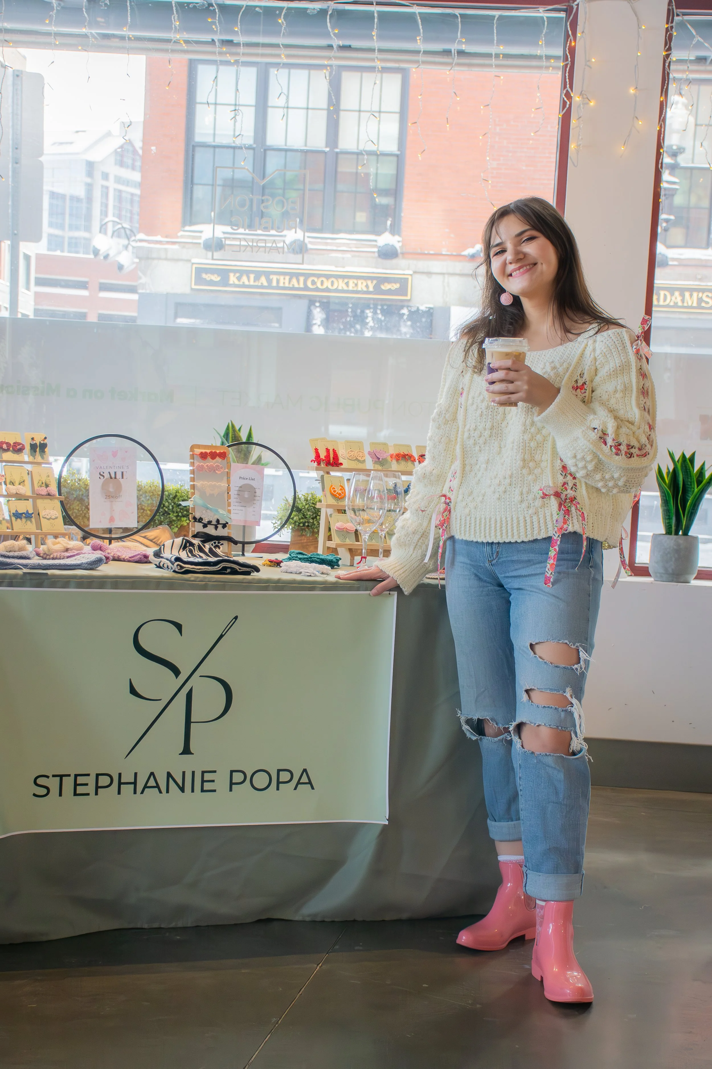 A young woman with brown hair, wearing a cream-colored sweater, ripped jeans, and pink rain boots, standing next to a table with a sign that reads 'Stephanie Popa'. She is smiling and holding a drink, inside a shop with a display of jewelry and acces