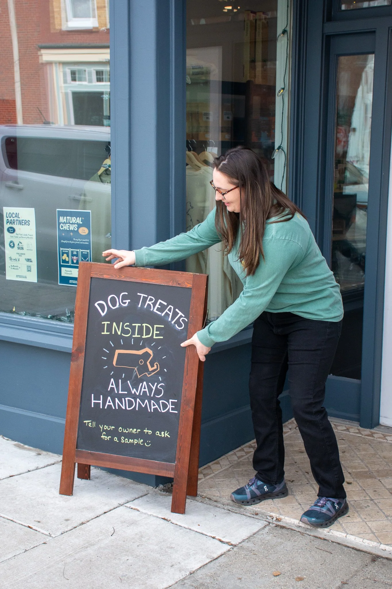 A woman in a green sweater and black pants adjusting a sidewalk chalkboard sign outside a store. The sign advertises that dog treats are inside and always handmade, with a note to tell the owner to ask for a sample.