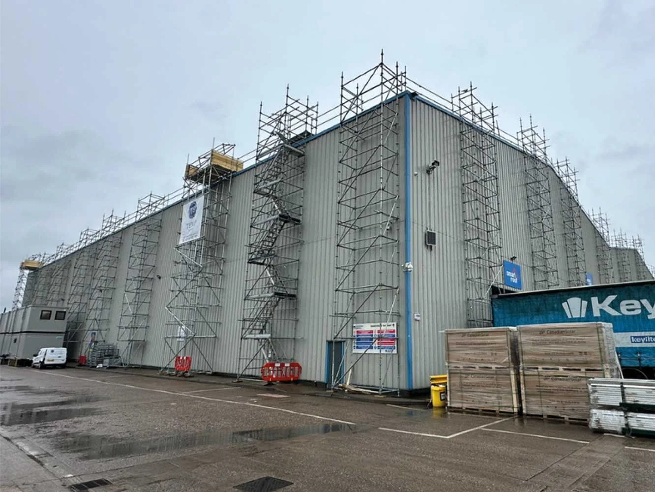 Large industrial warehouse under construction with scaffolding on the sides and construction materials and vehicles nearby, and a cloudy sky overhead.