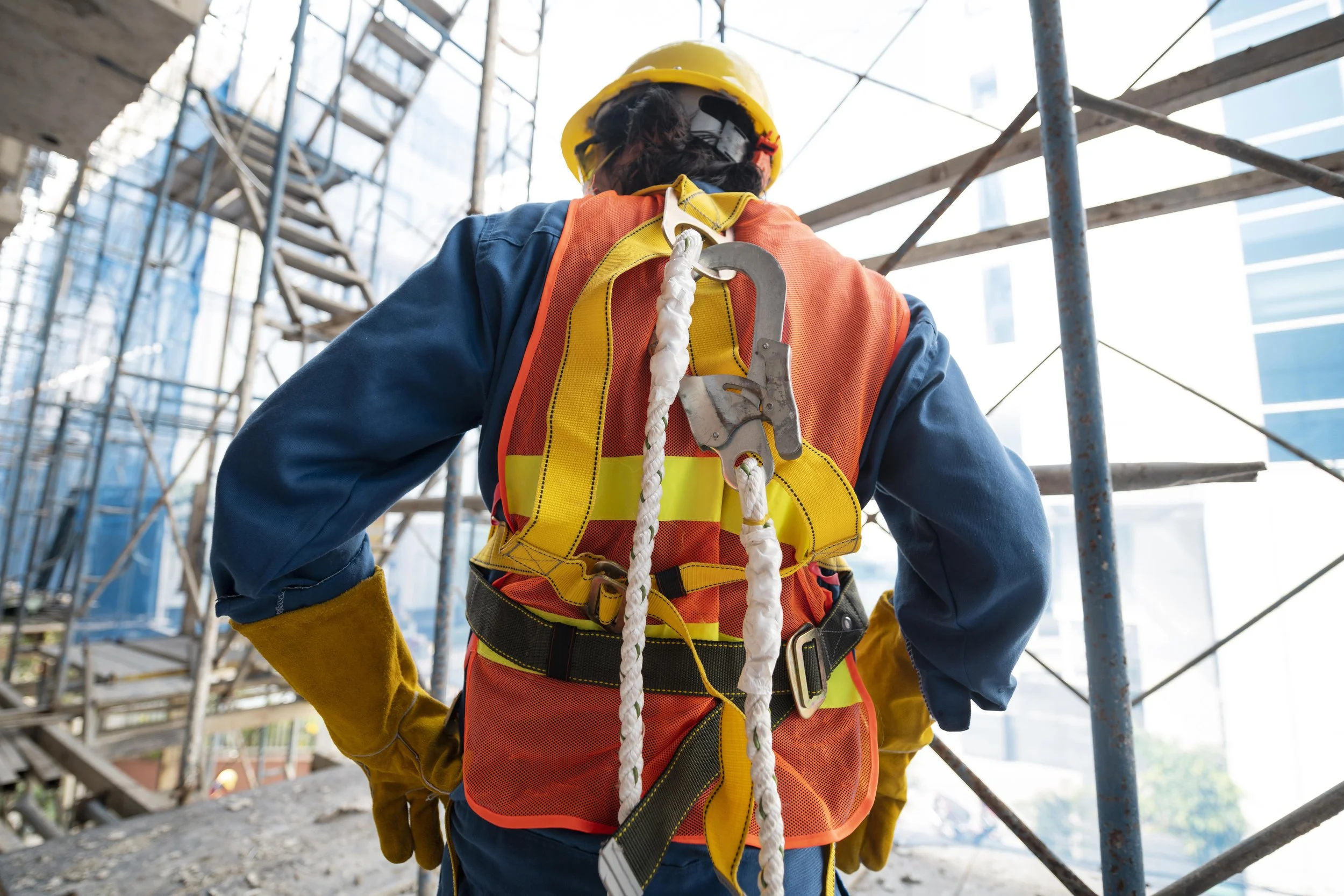 A construction worker wearing a yellow safety helmet, orange and yellow safety vest, blue long sleeve shirt, and yellow gloves, standing on a construction site surrounded by metal scaffolding.