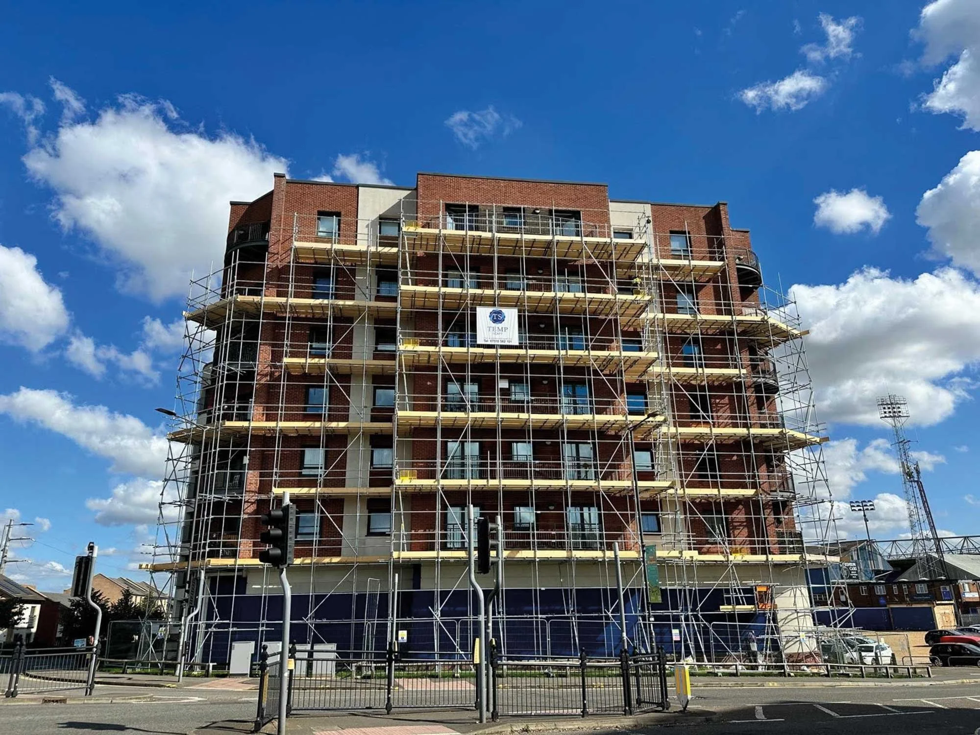A multi-story brick building under construction with scaffolding around it, set against a blue sky with scattered clouds.