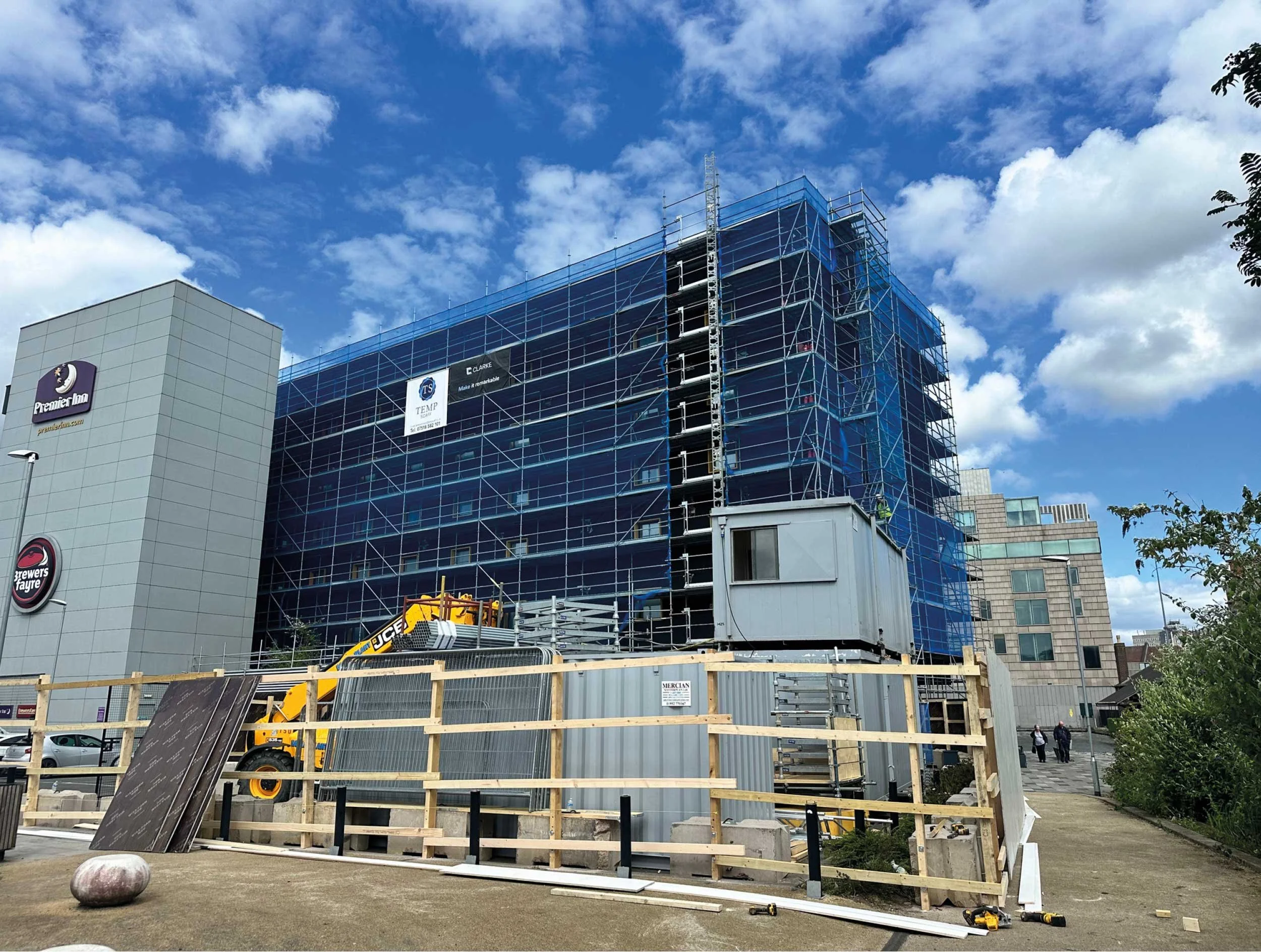 Under construction building with blue scaffolding, construction equipment, and a wooden fence in front, against a partly cloudy sky.