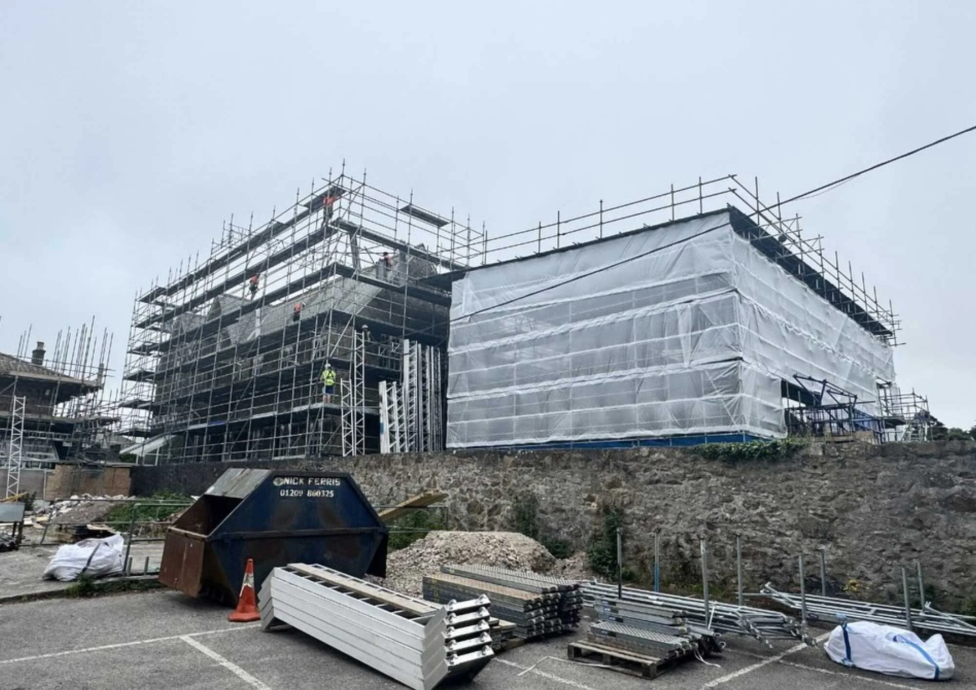Construction site with scaffolding around a building, some parts covered with white protective sheeting, construction materials and equipment on the ground, and a cloudy sky overhead.
