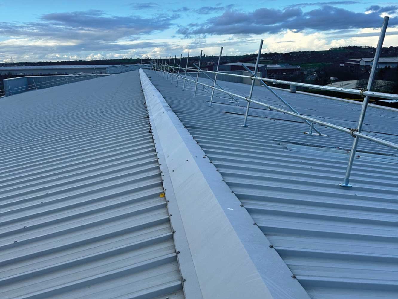 The image shows a metal rooftop with a central seam and protective railing. The sky is partly cloudy, and industrial or commercial buildings are visible in the background.