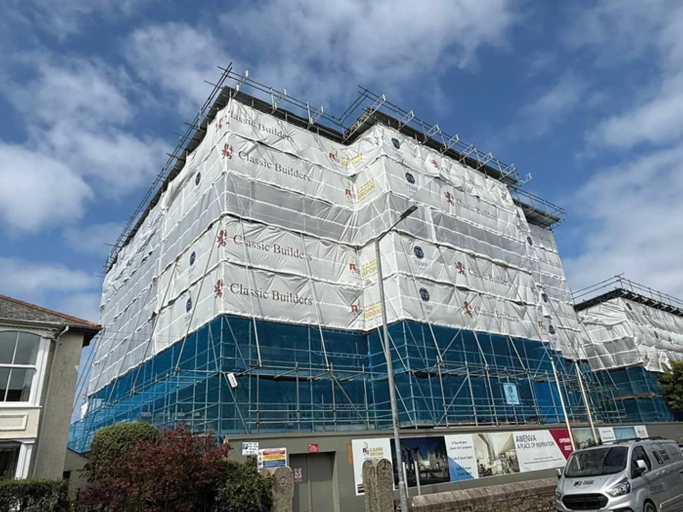 Building under construction wrapped in white and blue protective sheeting with scaffolding and a partly cloudy sky.