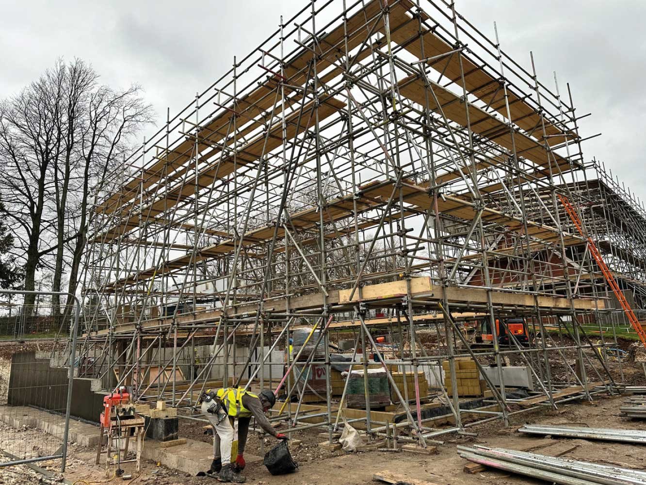 Construction site with scaffolding surrounding a building under construction, workers wearing safety gear, and construction materials scattered around on a cloudy day.