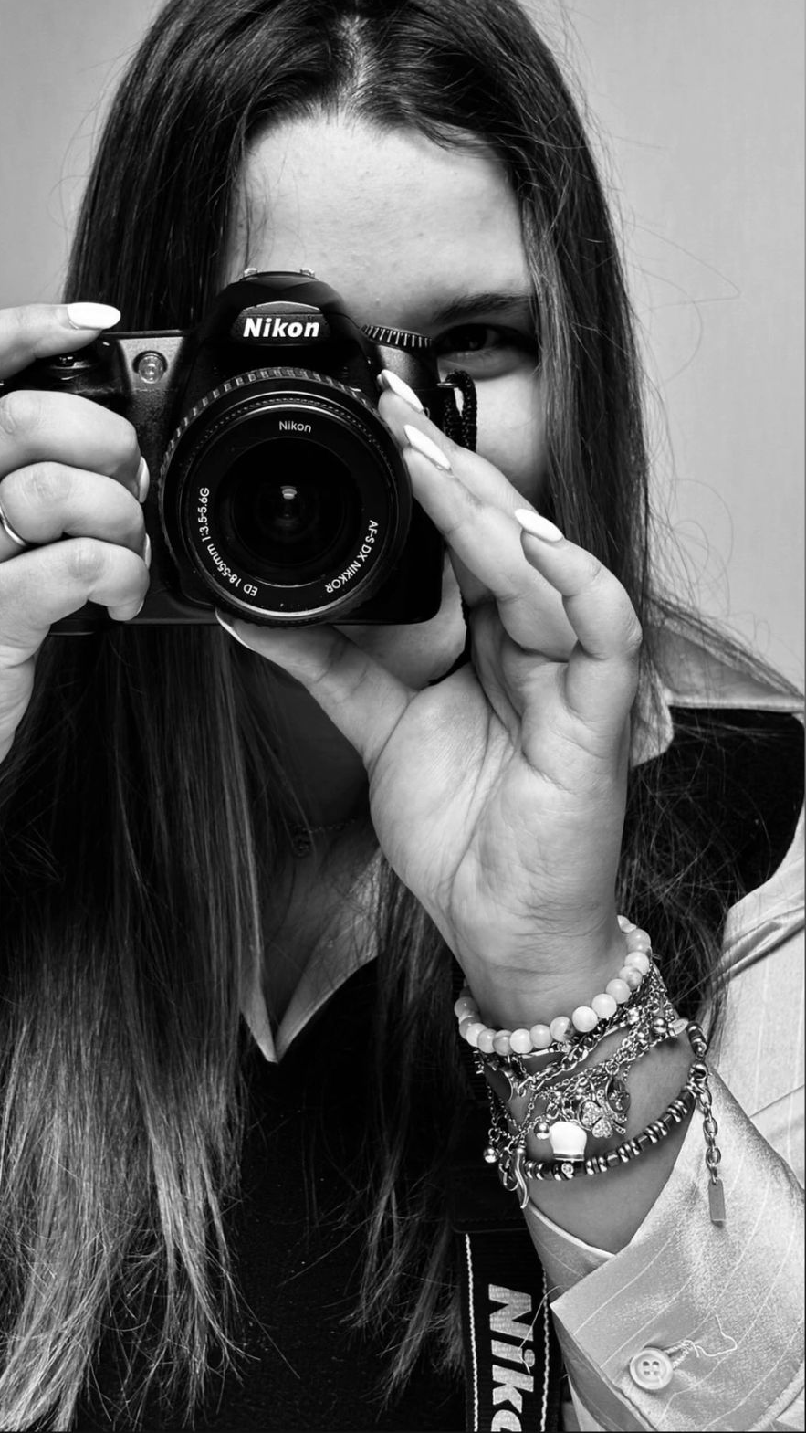 Una donna con lunghi capelli scuri, che prende una foto di se stessa tramite una fotocamera Nikon. Indossa vari braccialetti e ha un abito con colletto bianco.