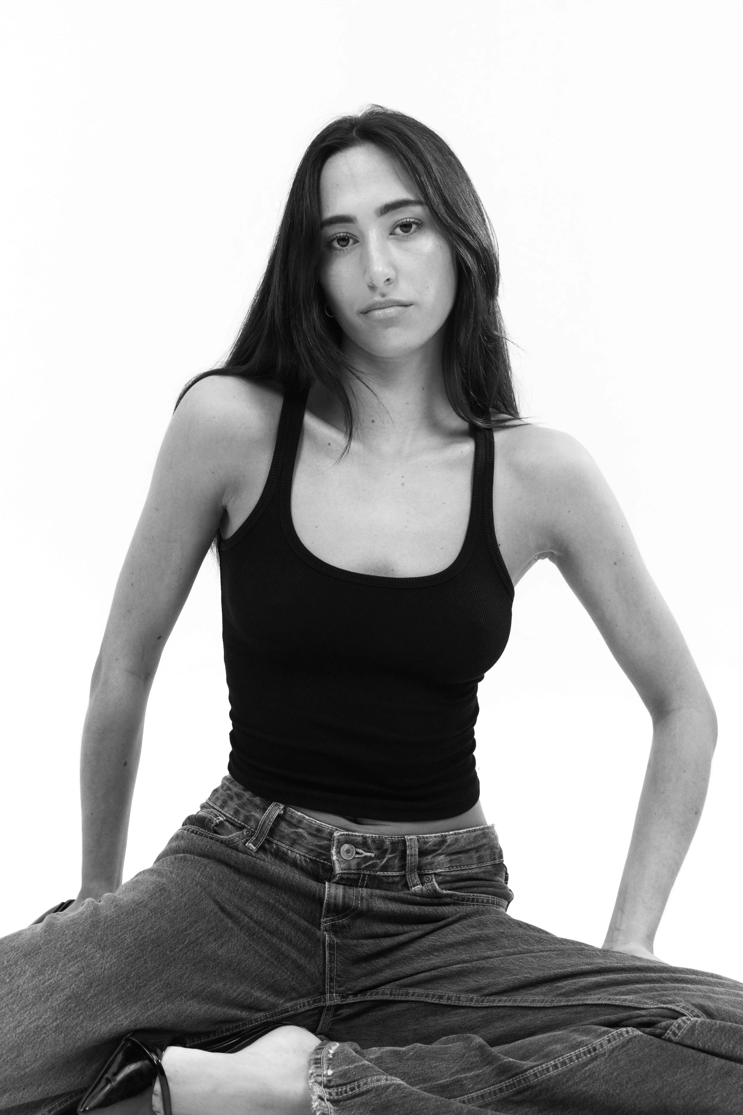 Black and white photo of a young woman with long hair, wearing a black tank top and jeans, sitting cross-legged against a plain background.