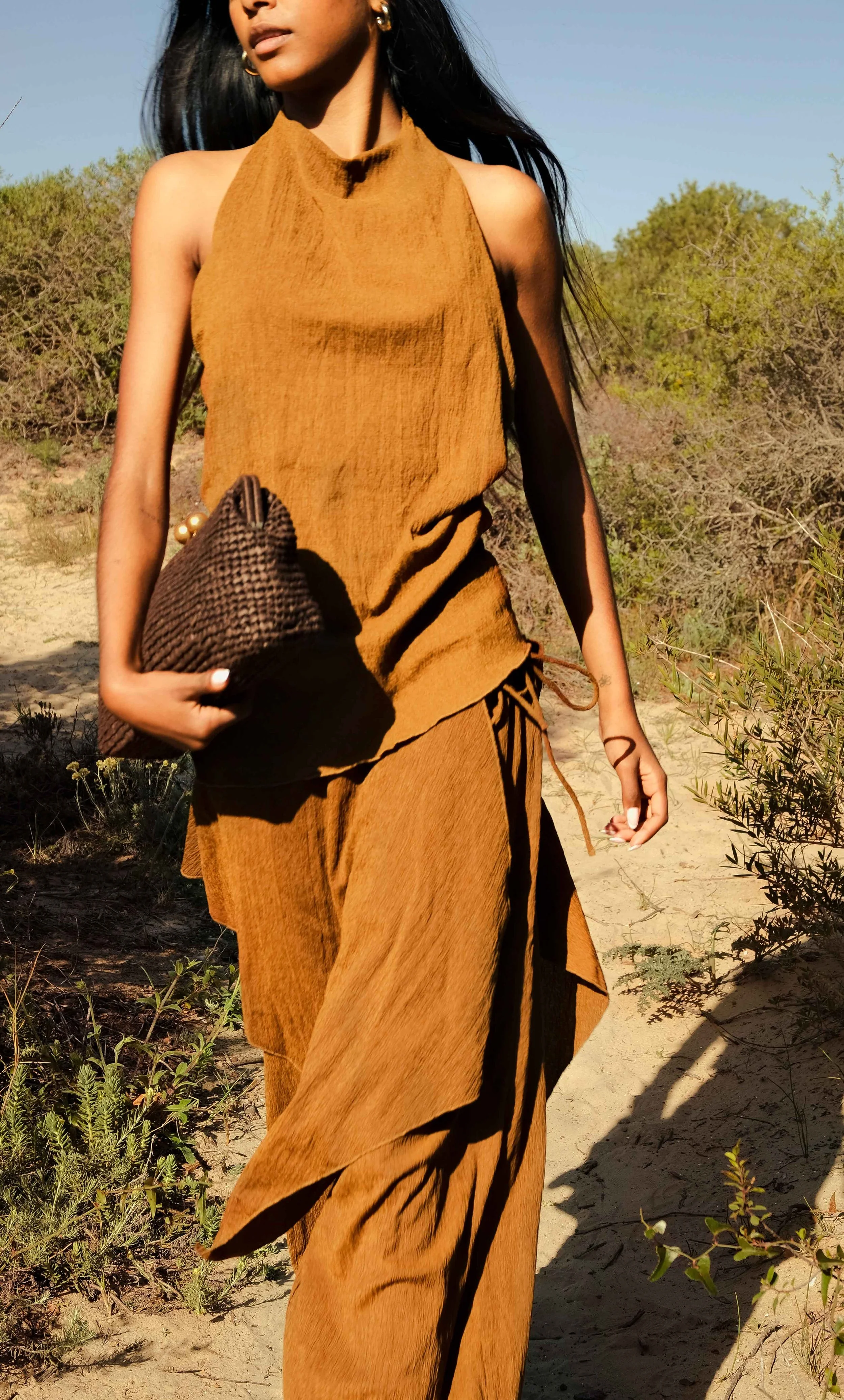 A woman wearing a brown sleeveless top and matching pants standing outdoors on a sunny day, holding a patterned brown clutch bag in her right hand. There is desert-like vegetation and clear blue sky in the background.