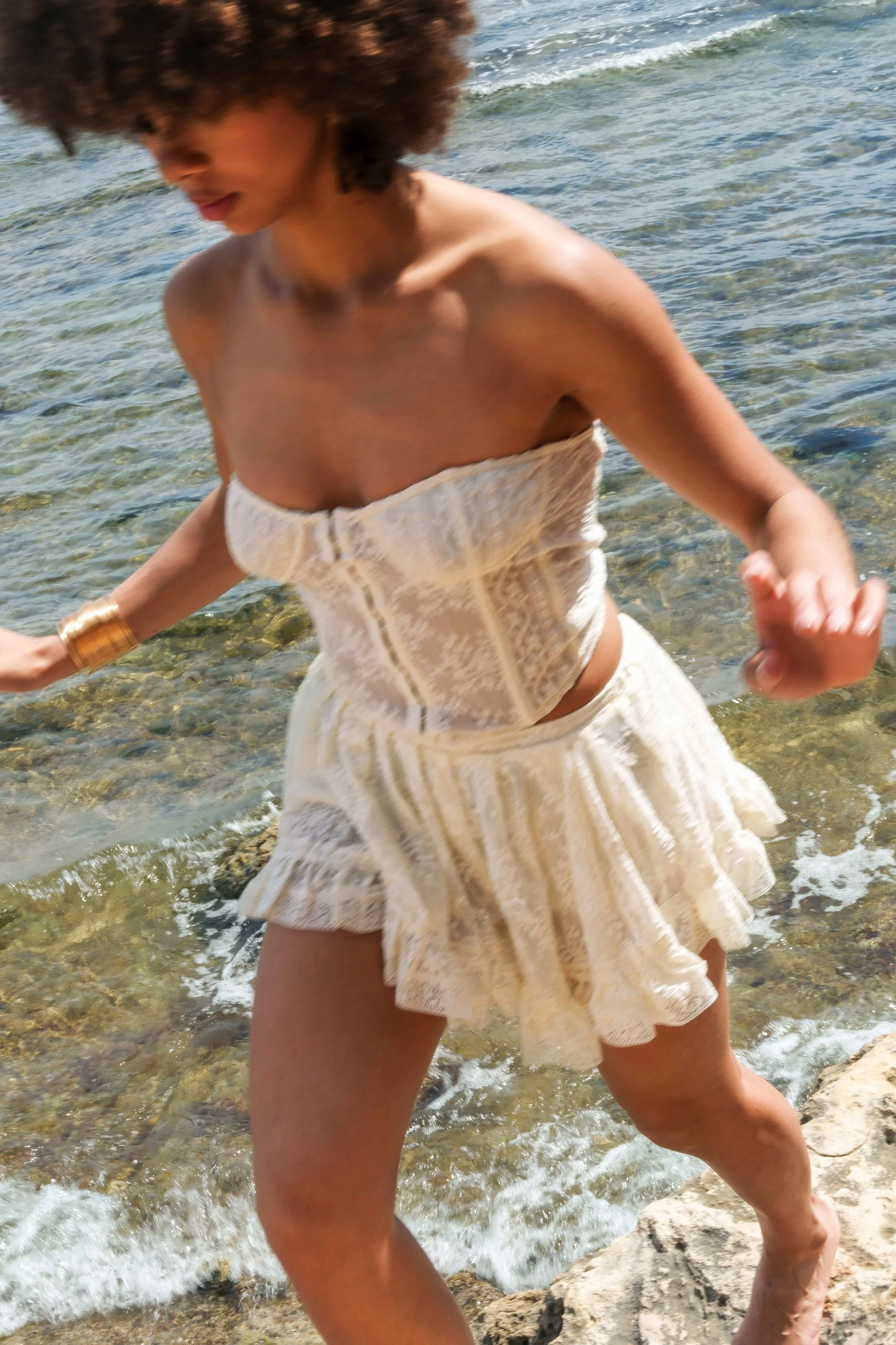 Woman wearing a white strapless dress with lace detail, walking by the water on a rocky shoreline.