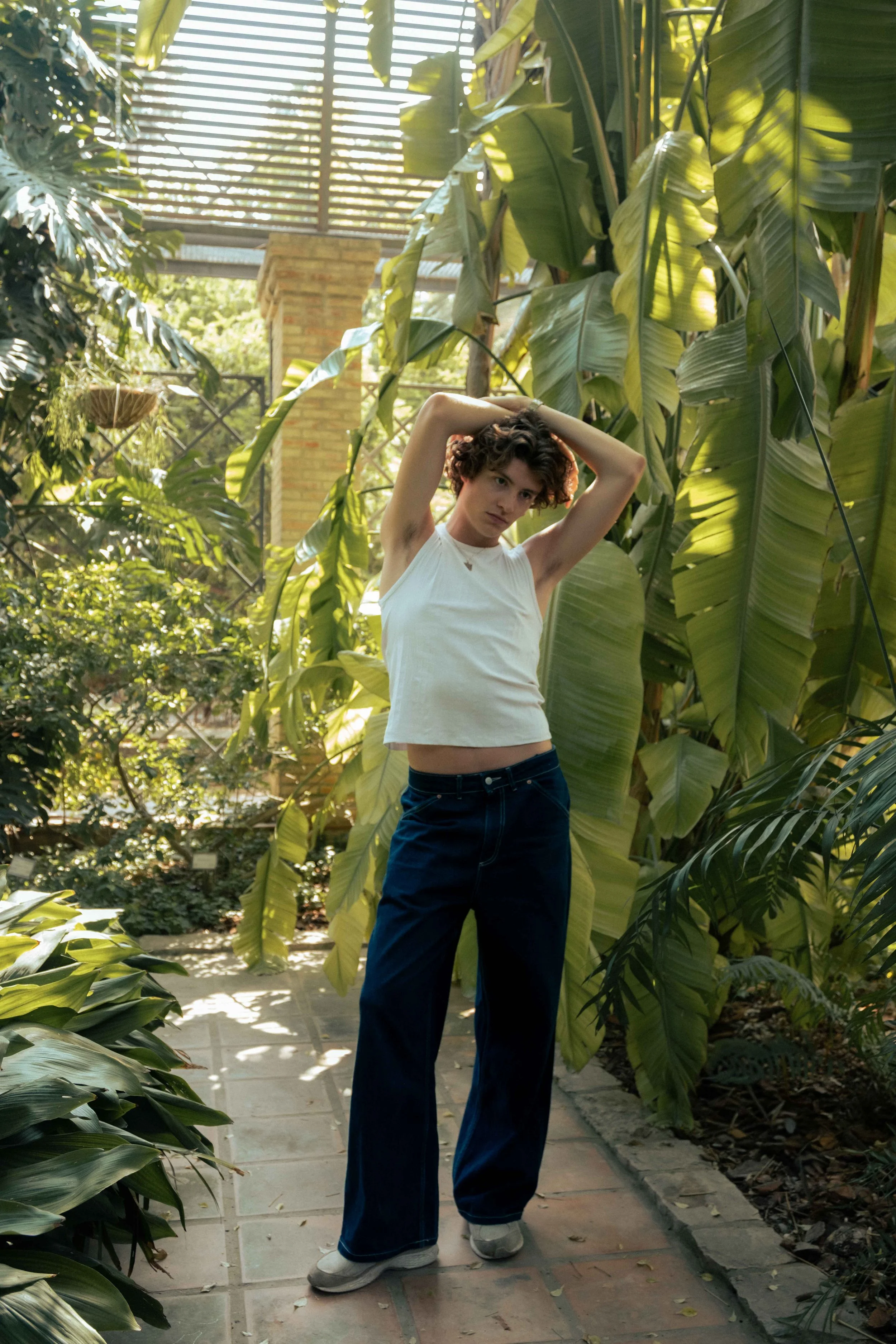 A young woman with short curly hair in a white sleeveless top and dark jeans stands on a stone pathway inside a lush green botanical garden with large tropical leaves around her.
