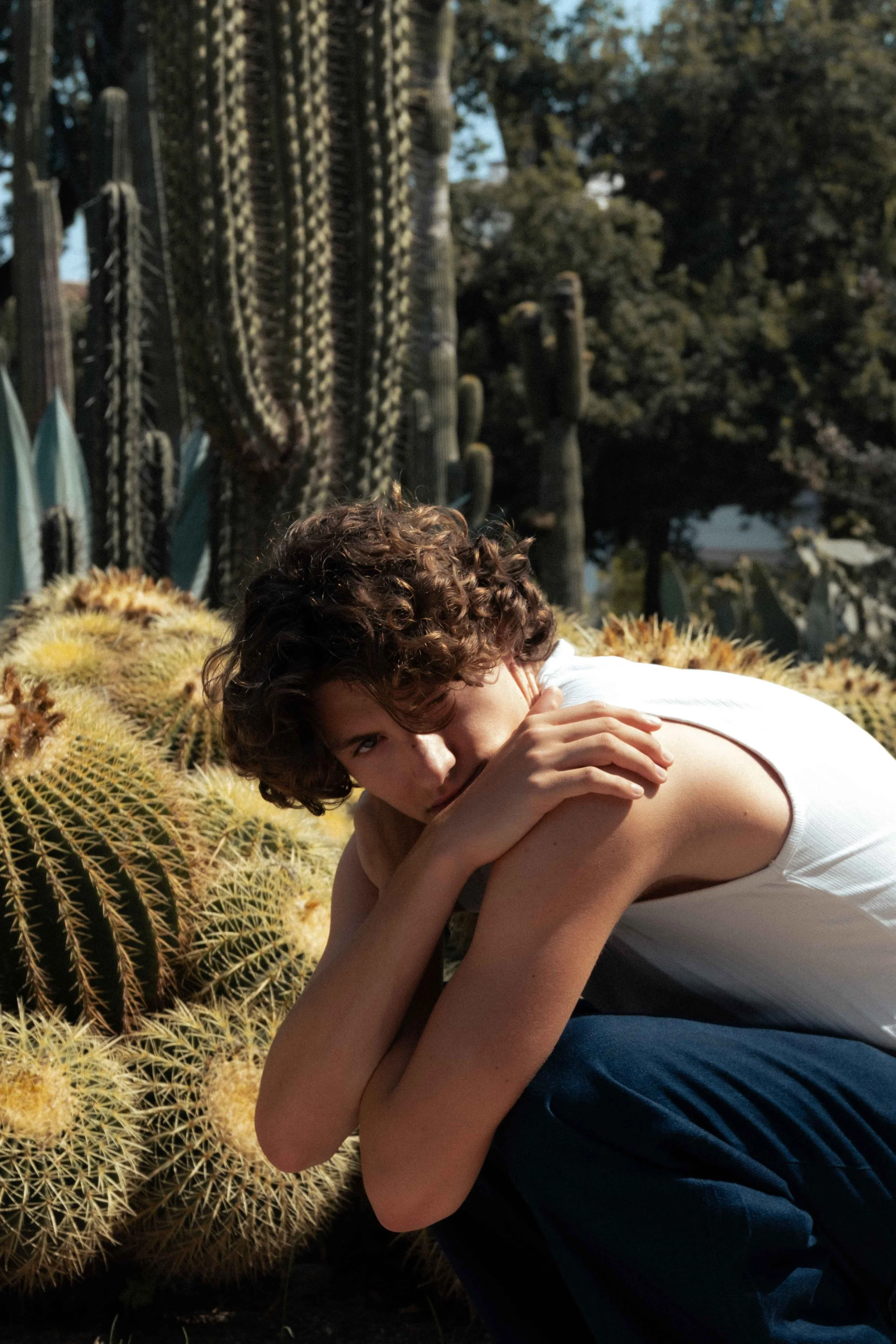 A young person with curly brown hair crouching next to a cluster of large, round golden barrel cacti outdoors, with tall cacti and trees in the background.