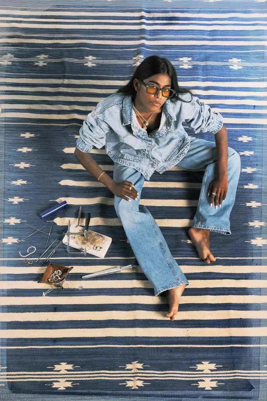 Person sitting on a blue and white patterned rug, surrounded by jewelry-making tools, wearing sunglasses, a denim jacket, and jeans.