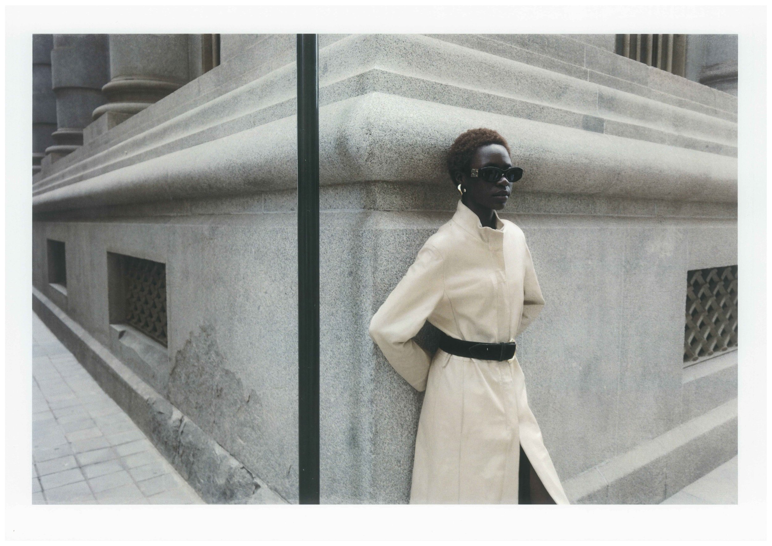 A woman with short hair wearing sunglasses and a beige trench coat with a black belt, standing against a stone building with architectural details, on a city sidewalk.